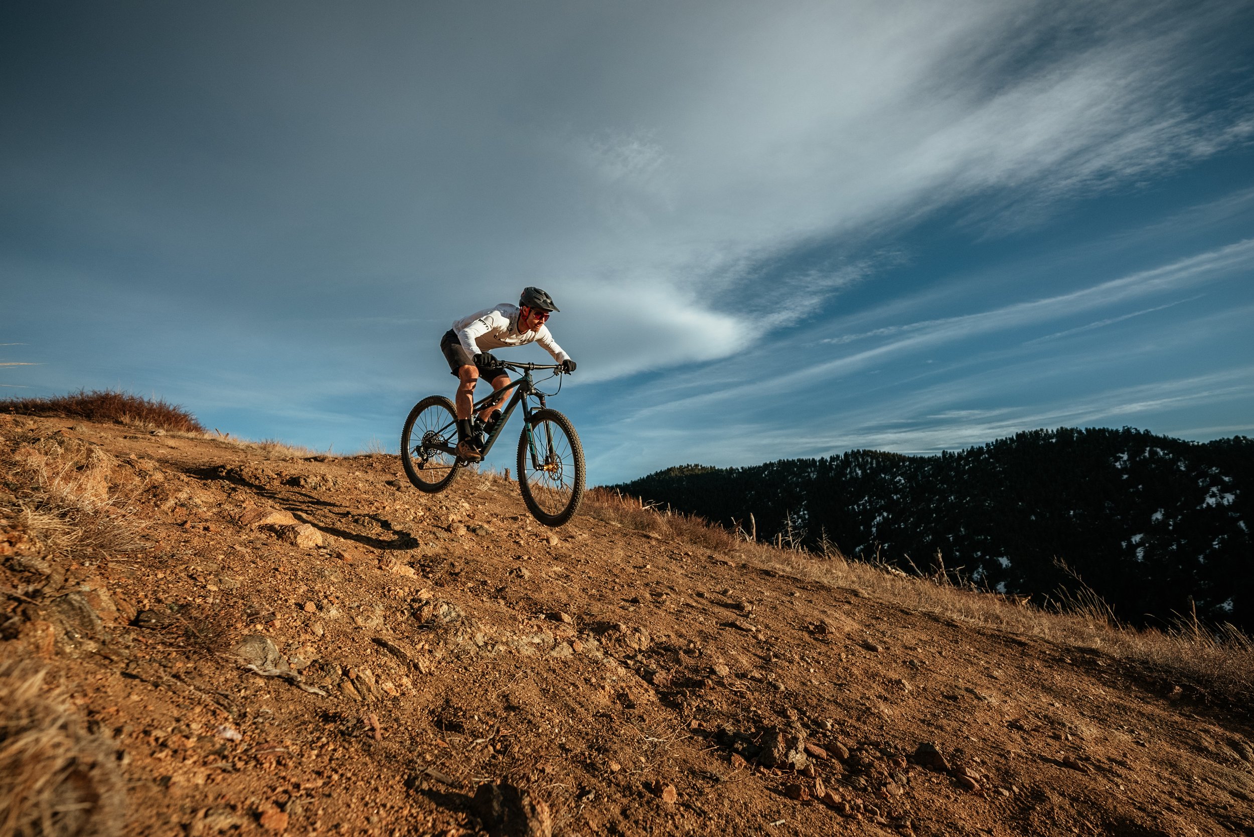 A person wearing a helmet and white long-sleeve shirt riding a mountain bike downhill on a dirt trail with a mountainous landscape and blue sky in the background.