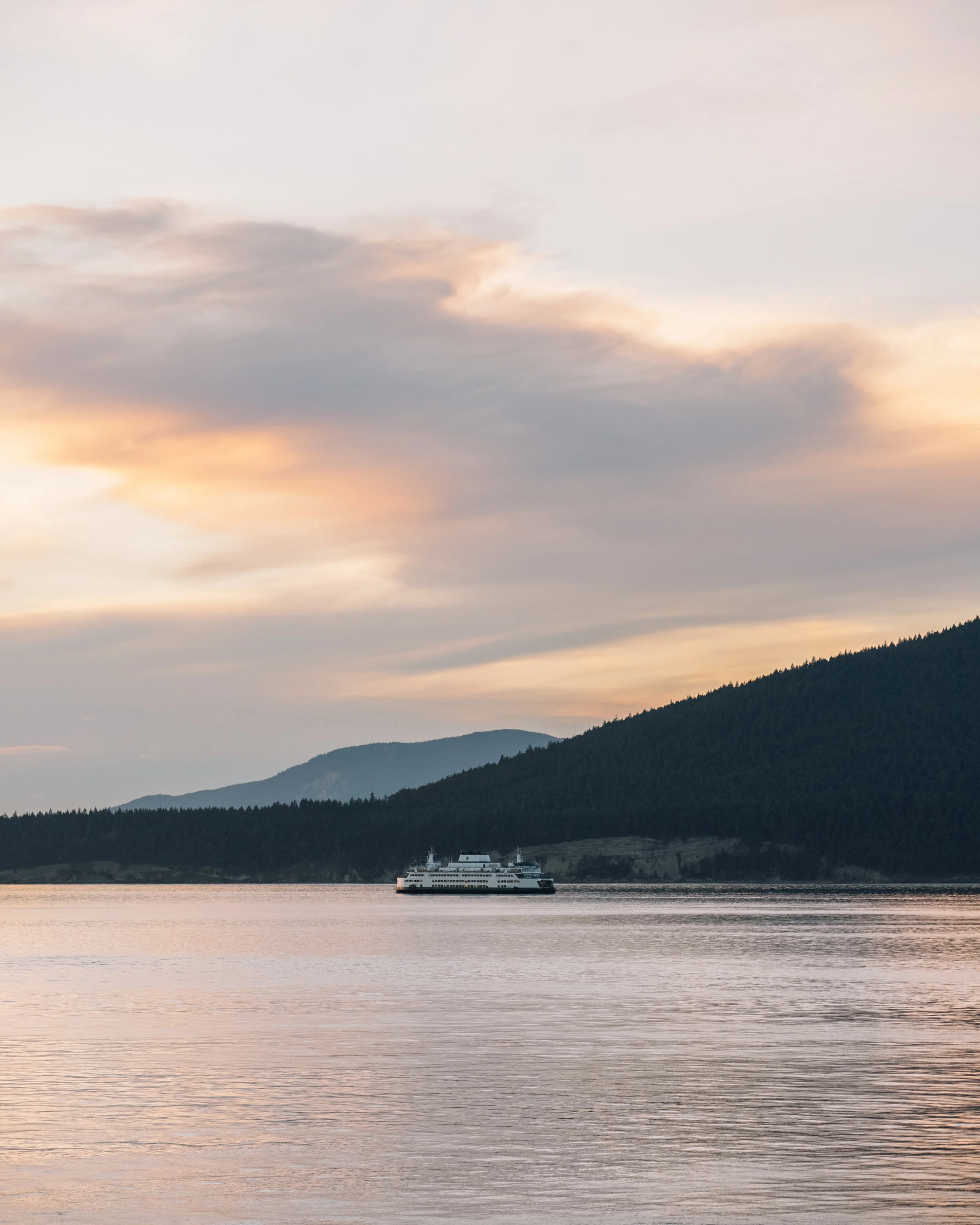 A large ferry boat sailing on calm water with a backdrop of forested mountains and a colorful sky at sunset.