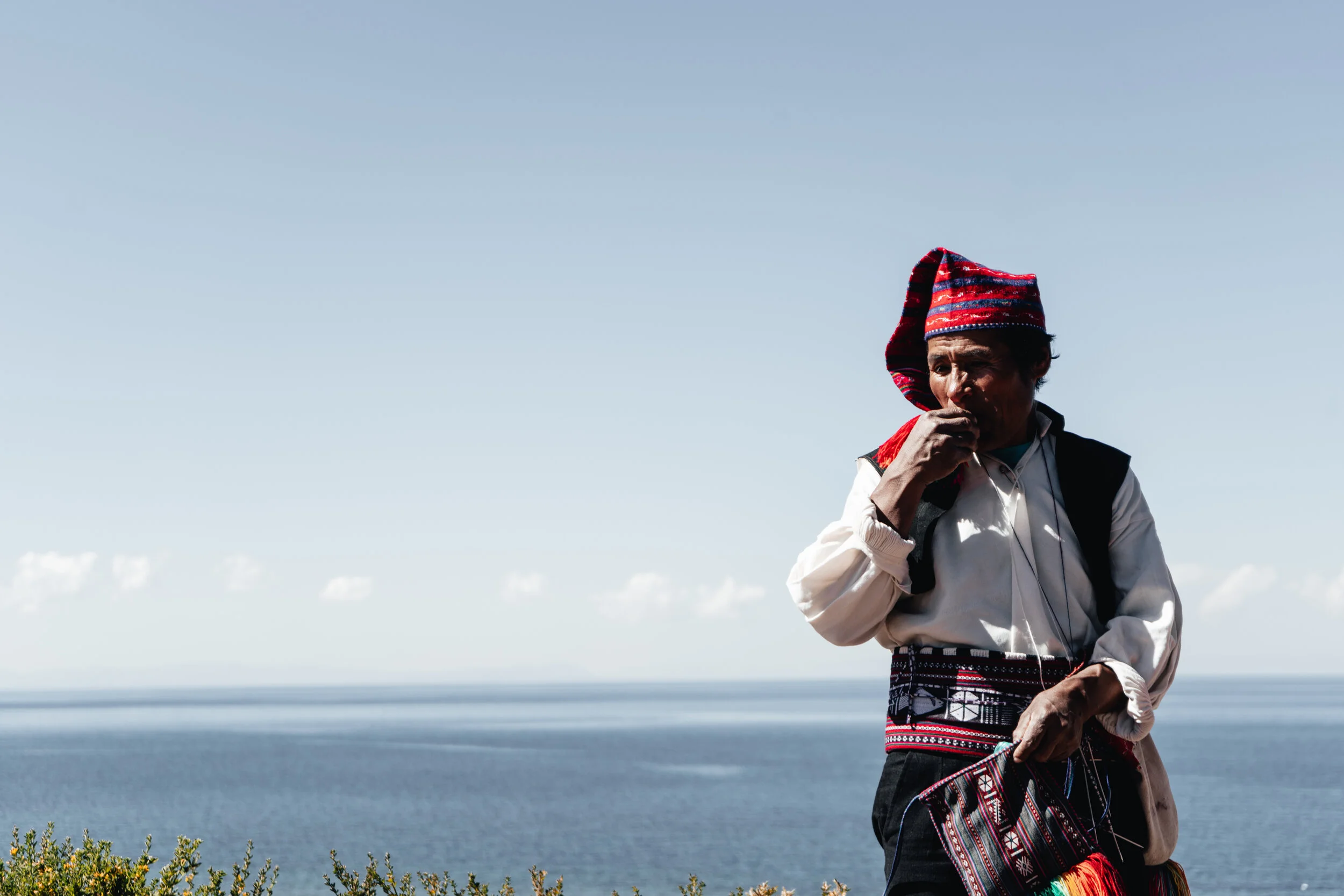 An elderly man wearing traditional clothing, including a red and black embroidered hat and sash, standing outdoors near the ocean with a clear sky in the background.