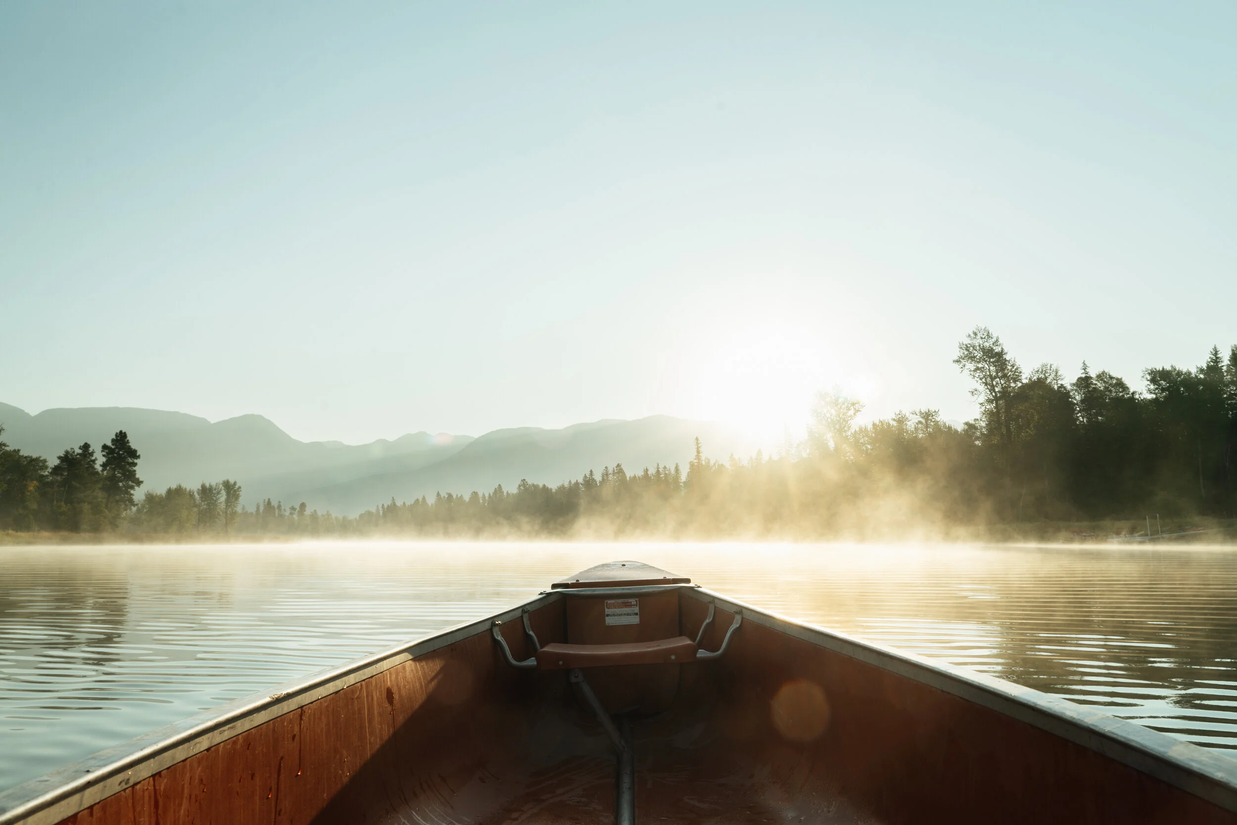 View from a boat on a calm lake at sunrise, with mist rising from the water and mountains in the background.