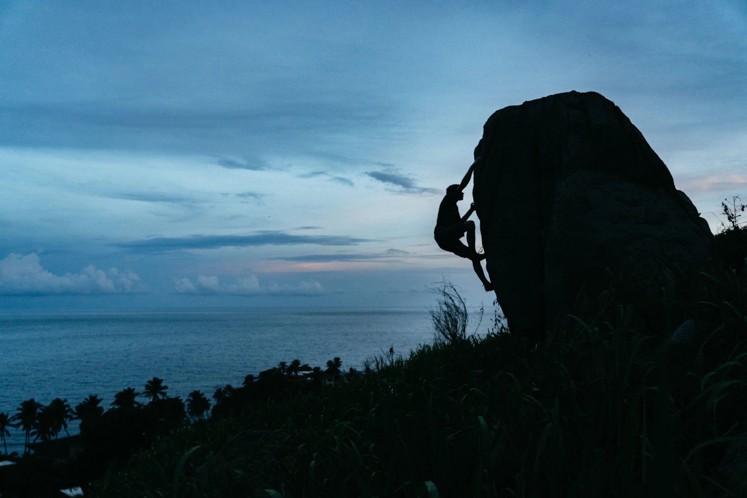 A person climbing a large rock in silhouette against a twilight sky with an ocean and palm trees in the background.