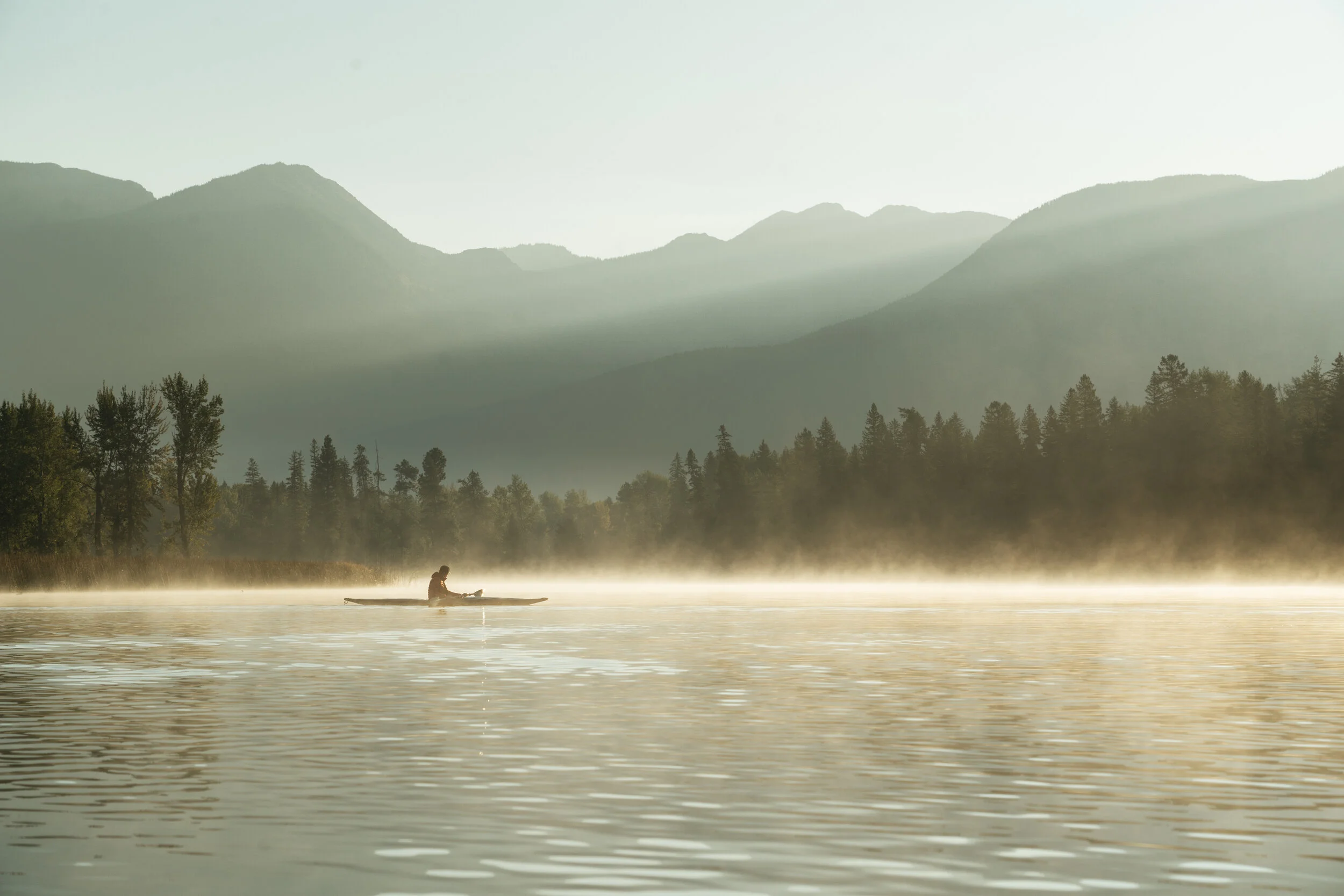 Person kayaking on a calm river during early morning with mist, trees along the shore, and mountains in the background.