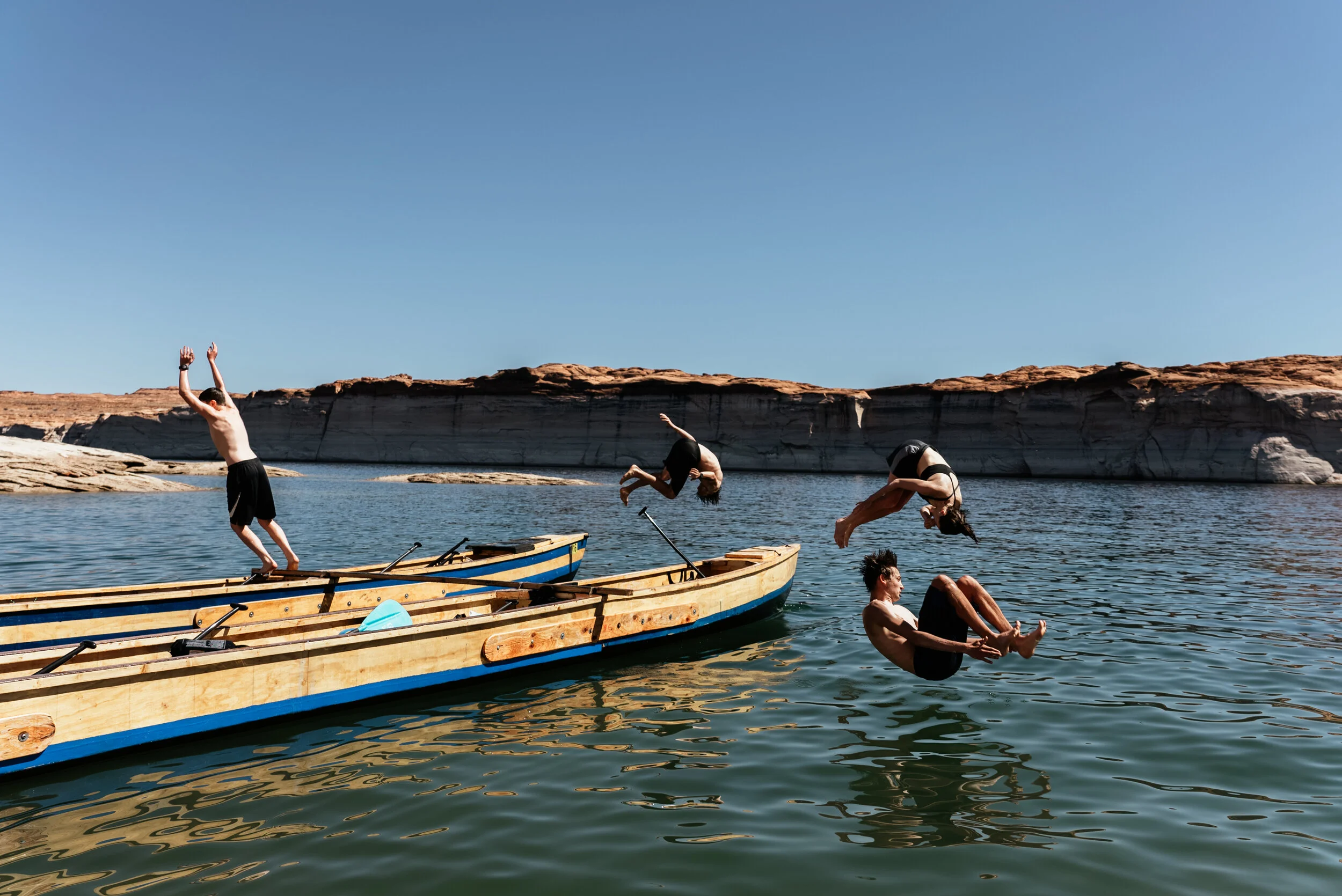 Four people jumping into water from wooden boats in a desert landscape with cliffs and clear sky.