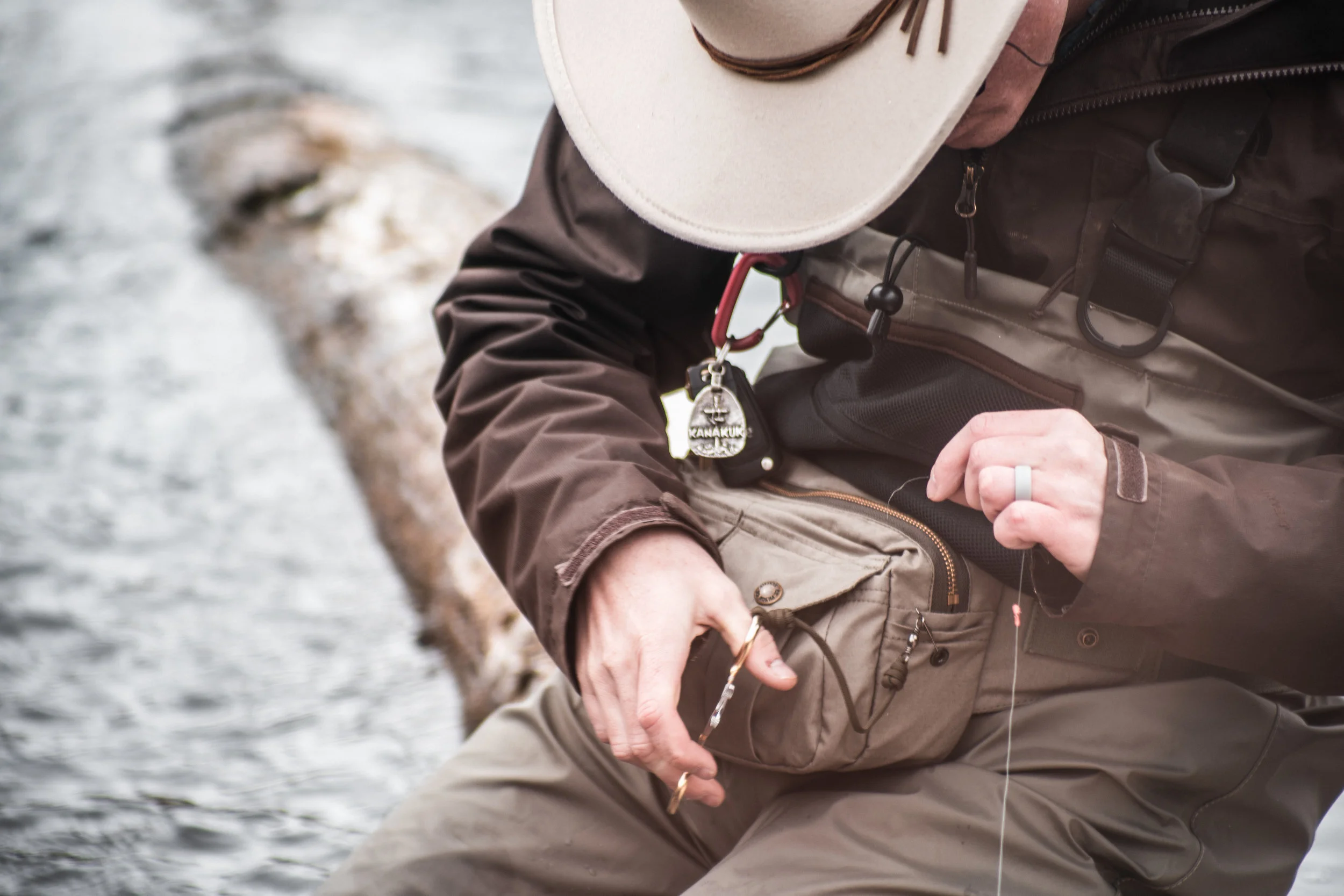 Person fishing near water, wearing a beige hat, brown jacket, and carrying a beige bag, with a visible fishing hook and a dog tag attached to the jacket.
