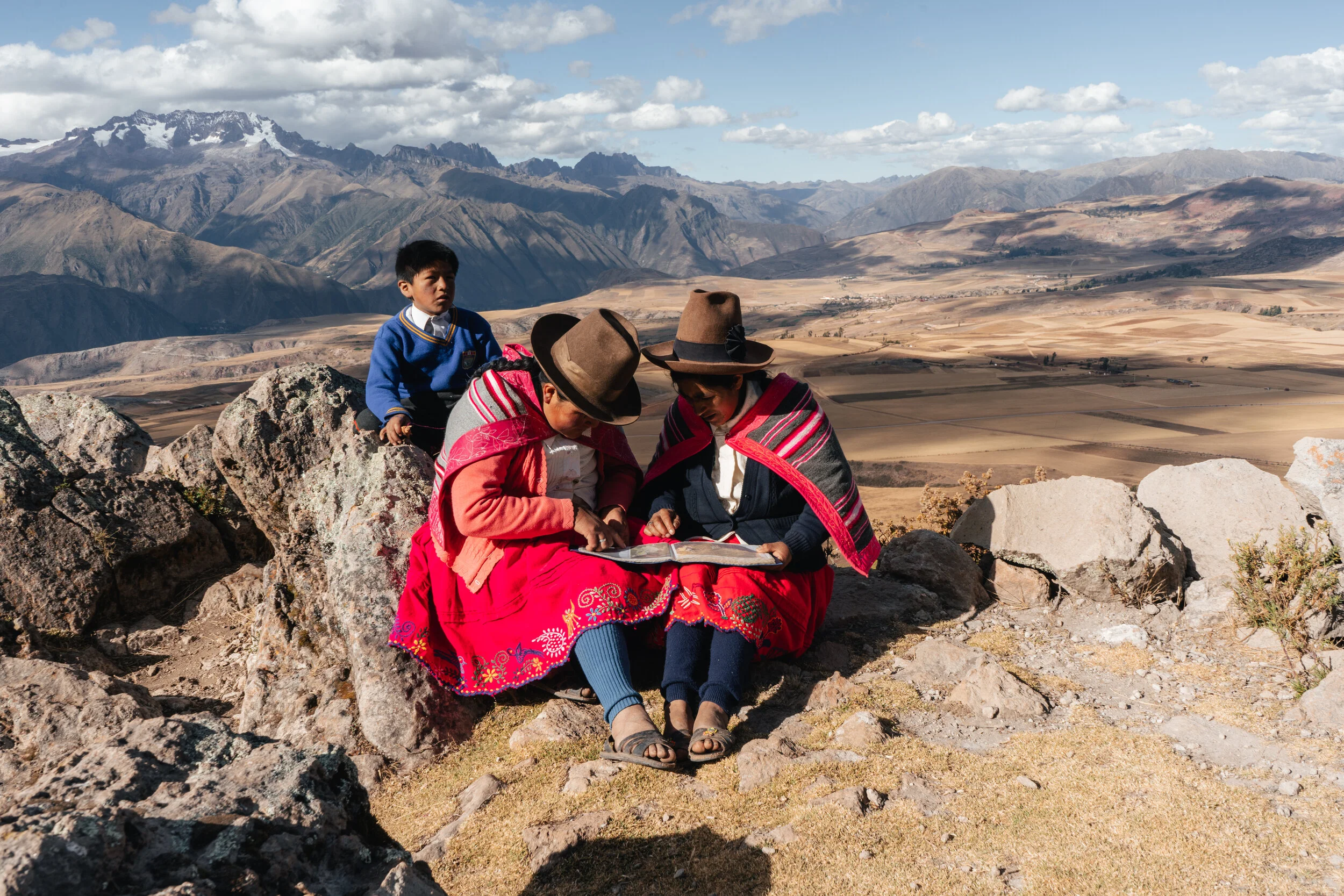 Two women wearing traditional Andean clothing and hats sitting on rocks, looking at a book or paper, with a young boy standing nearby behind them. They are on a hillside overlooking a valley with mountains and fields.
