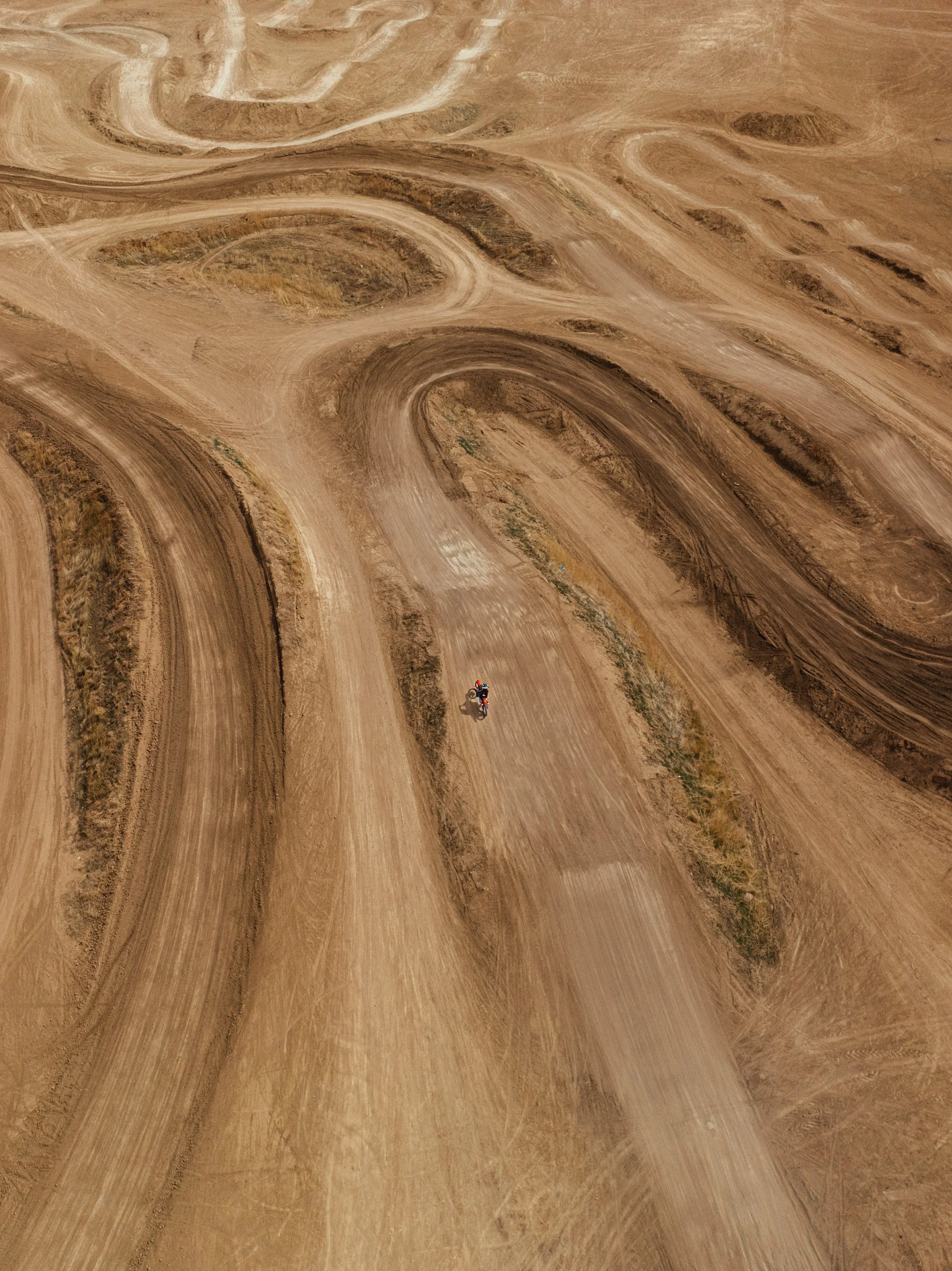 An aerial view of a dirt motocross track with multiple winding and curved paths, and a motorcyclist riding on one of the tracks.
