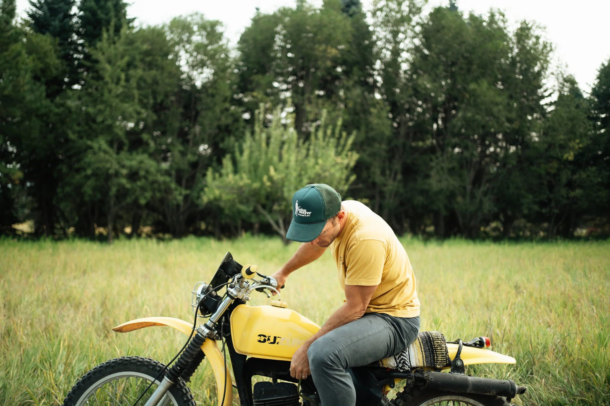 A man in a yellow t-shirt and gray pants sitting on a yellow Suzuki motorcycle in a grassy field, wearing a green cap, with lush trees in the background.