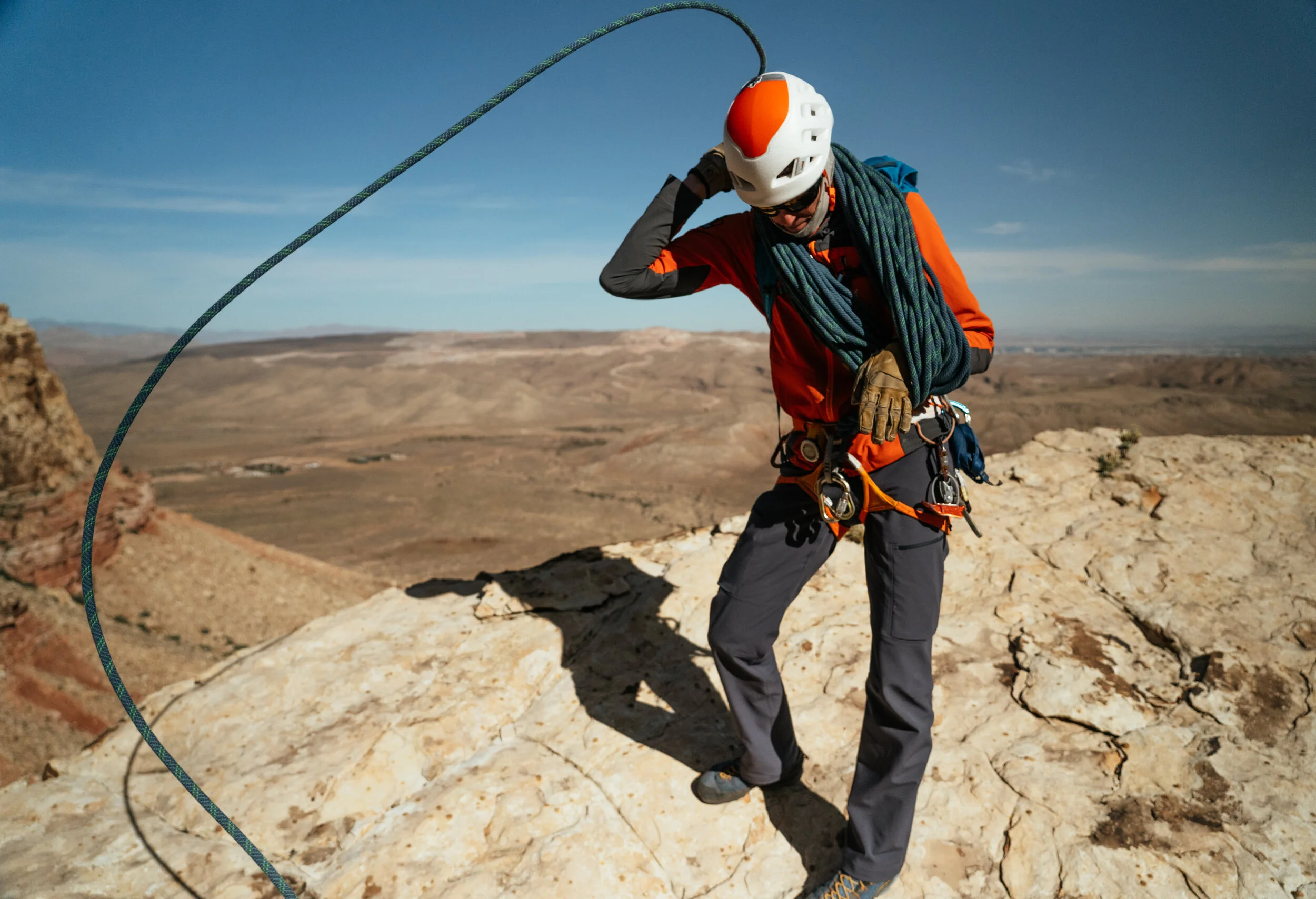 A person in outdoor climbing gear standing on rocky terrain, adjusting a climbing helmet, with climbed rope around their neck, in a desert landscape under a clear blue sky.
