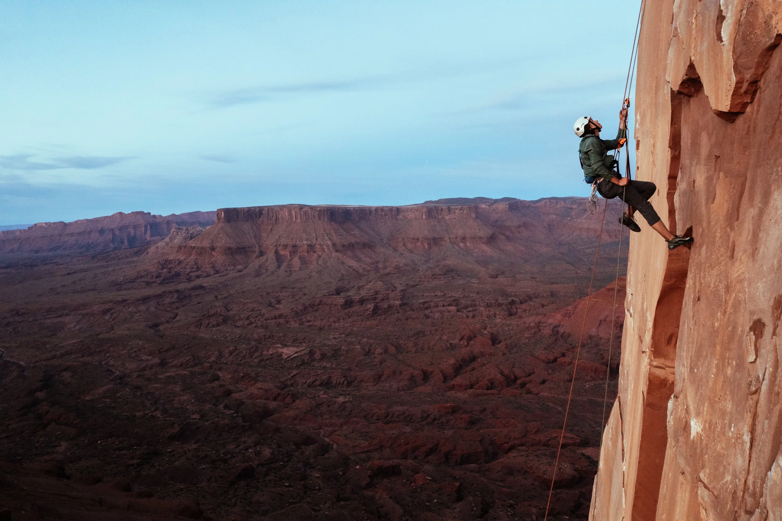 A person wearing a helmet and harness rock climbing on a cliff face with a mountainous desert landscape in the background during twilight.
