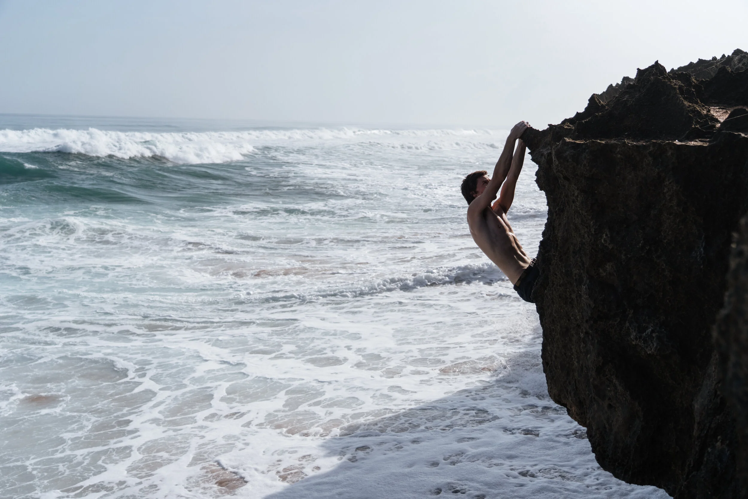 A man climbing a large rock on the beach near the ocean with waves in the background.
