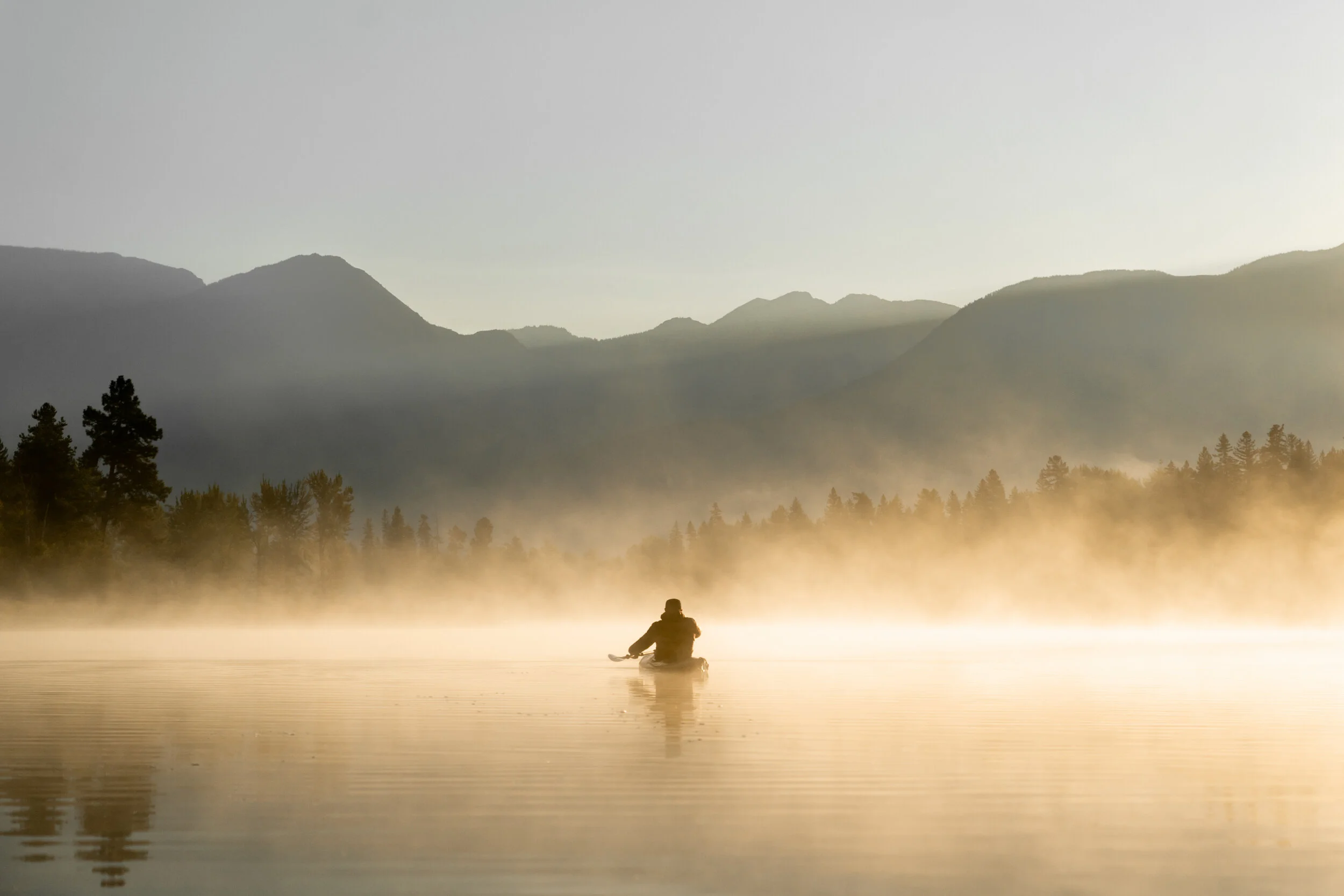 A person sitting in a kayak on a foggy lake with mountains in the background at sunrise or sunset.