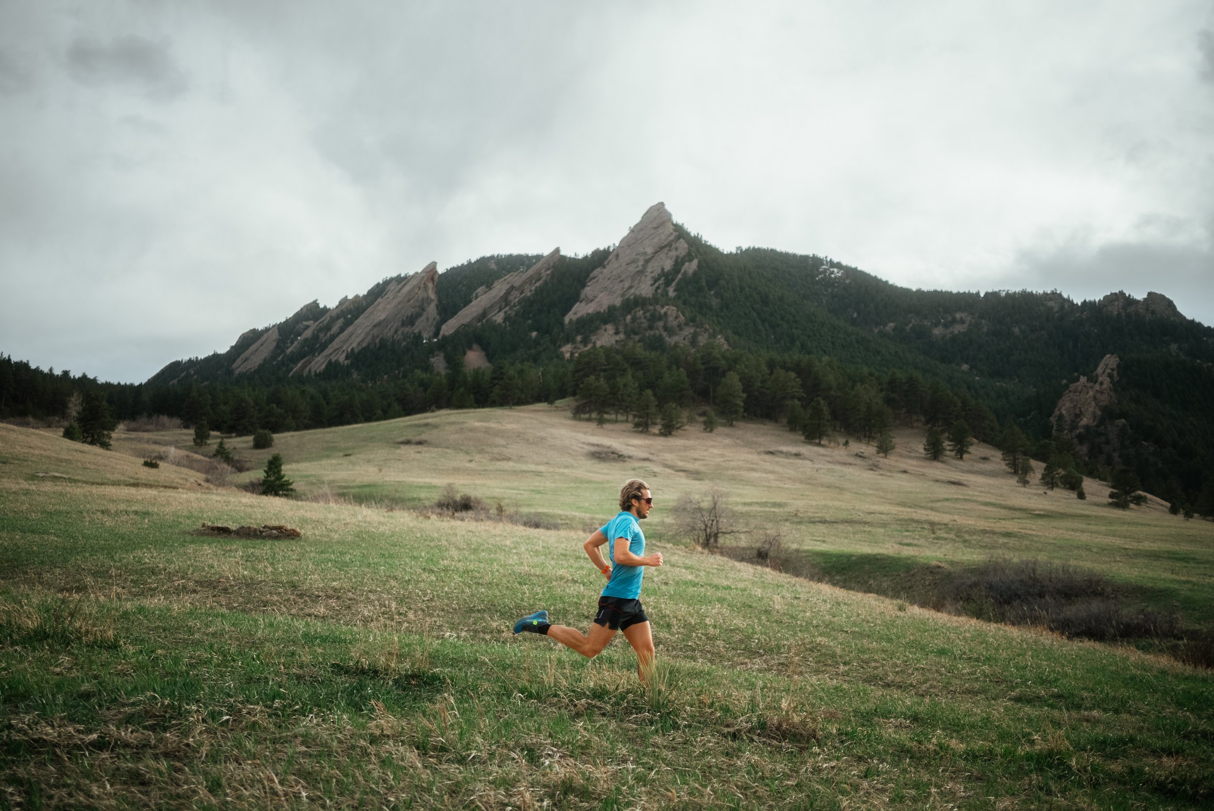A man running on a grassy field with rocky mountains and a cloudy sky in the background.