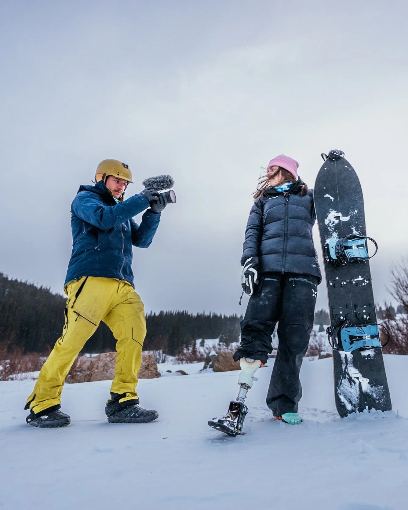 A person with a prosthetic leg stands in the snow with a snowboard against their leg, while another person records them with a camera during winter outdoor activity.