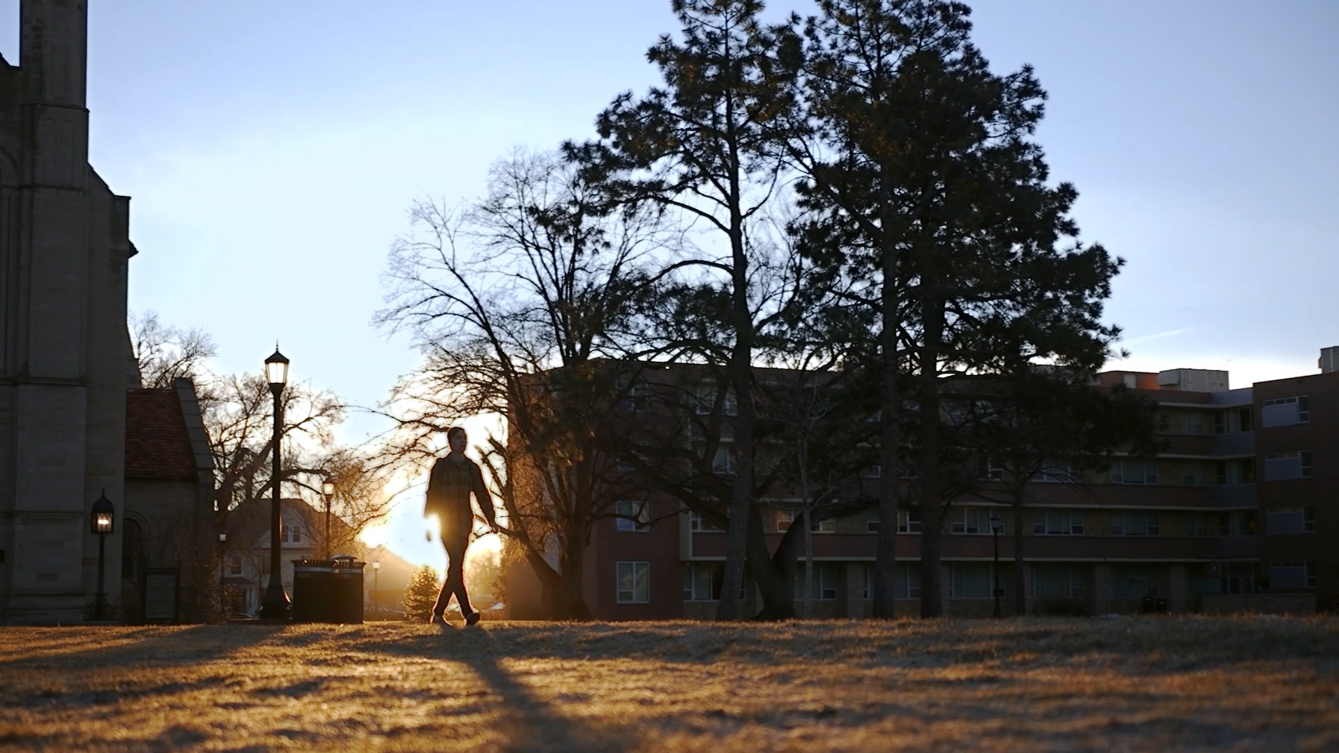 A person walking outdoors at sunset with trees and buildings in the background.