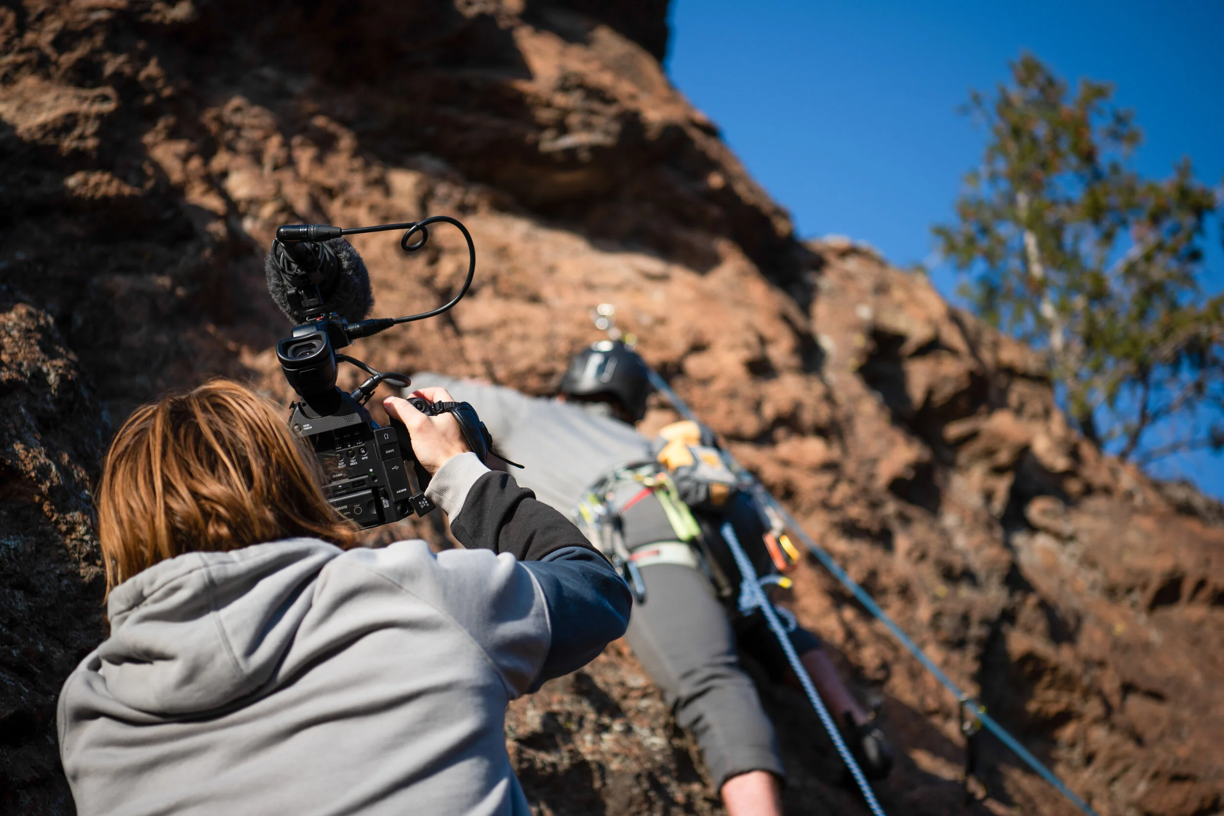 A person is filming a climber ascending a rocky cliff with a professional camera, while the climber is using safety gear and ropes.