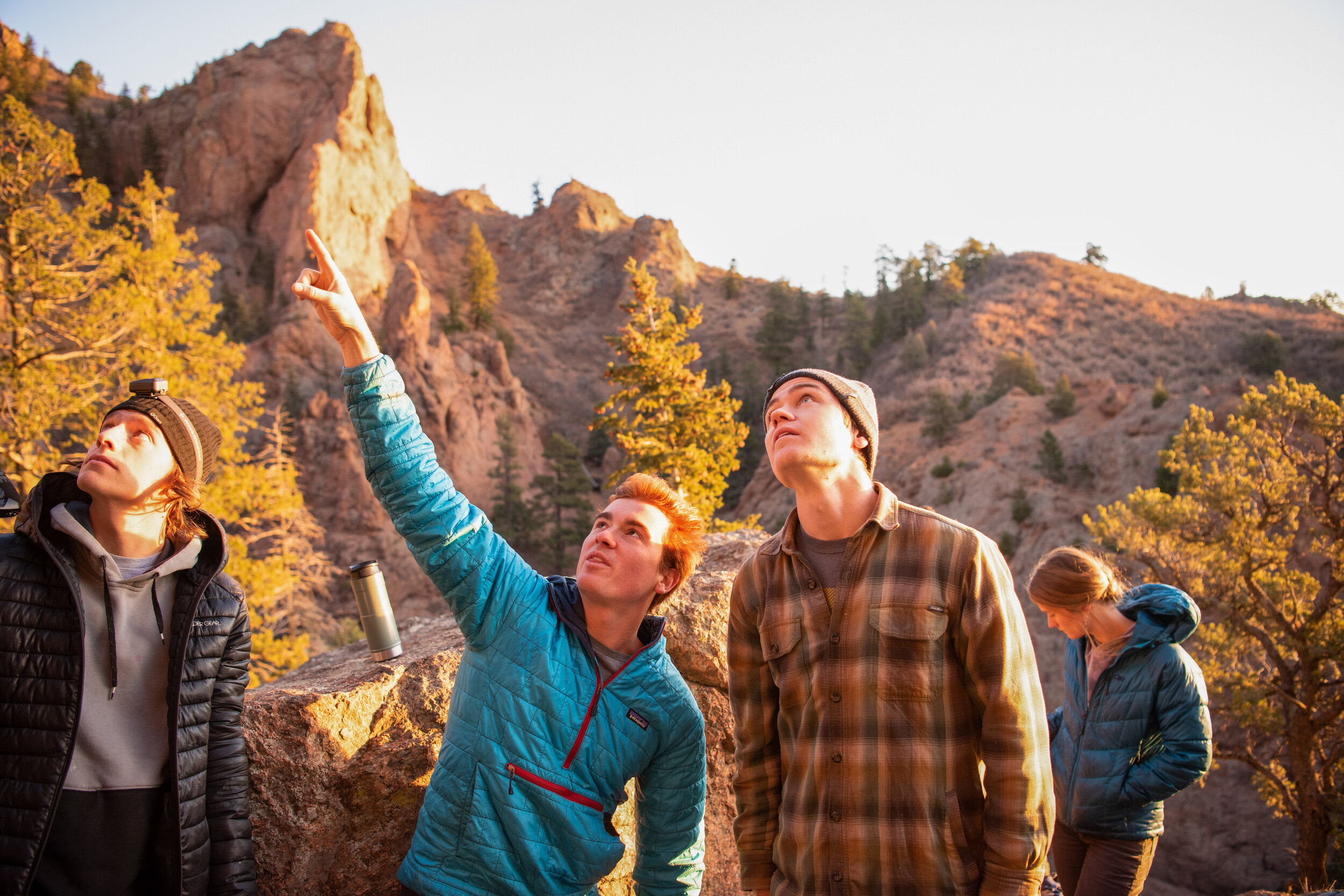Four young people in outdoor jackets and hats, standing on a rocky trail in a mountainous area with trees, during the golden hour, looking up at the sky. One person is pointing upwards, possibly at something in the sky.