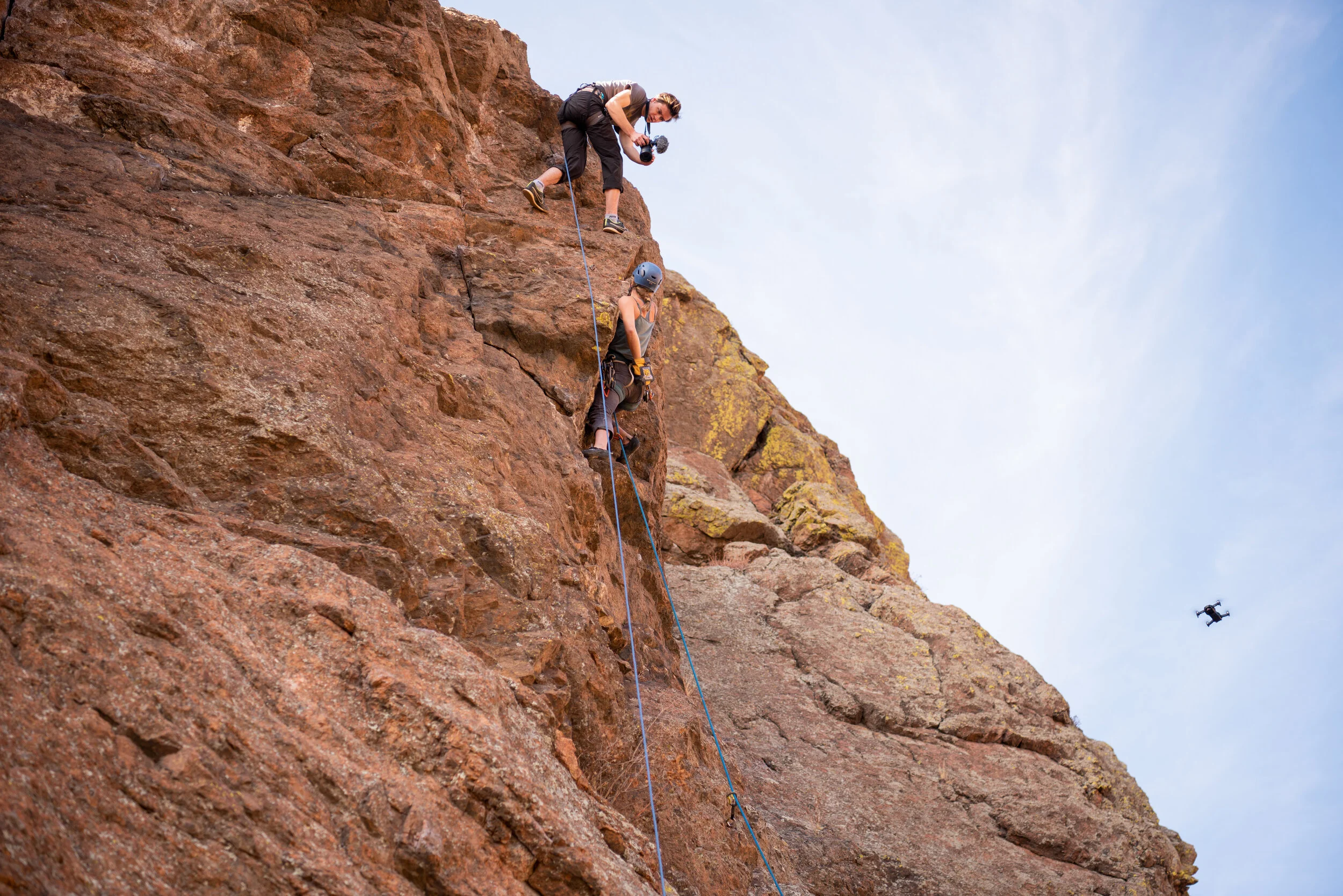Two rock climbers on a steep red rock face, one wearing a helmet and harness, assisted by a photographer holding a camera and standing above them. A drone flies nearby against a cloudy sky.
