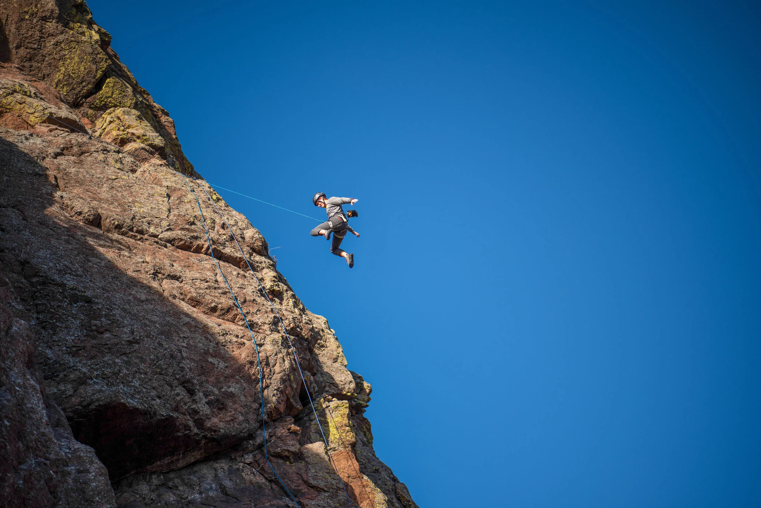 A person rock climbing on a steep cliff, wearing a helmet and harness, mid-air against a clear blue sky.