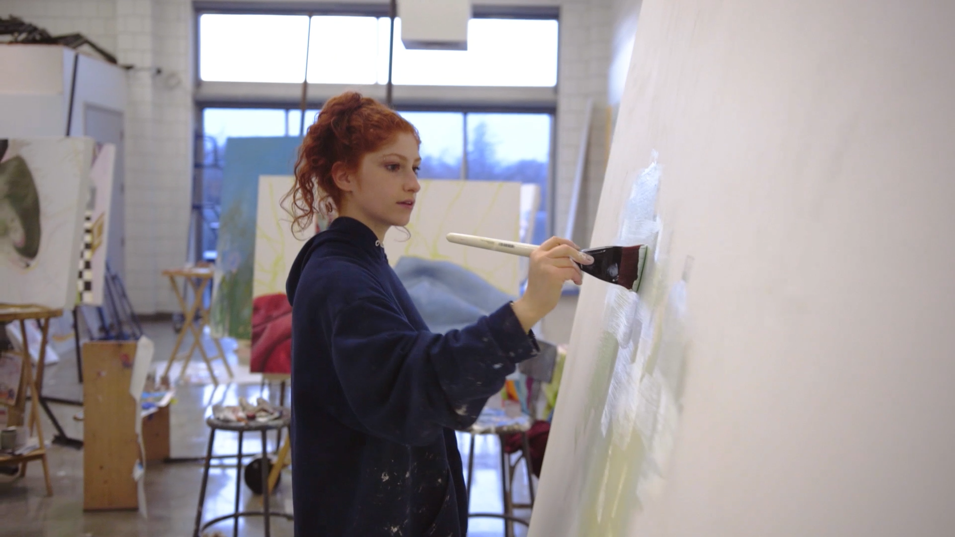 A young woman with red curly hair painting on a large canvas in an art studio, wearing a dark hoodie with paint stains, and holding a paintbrush.