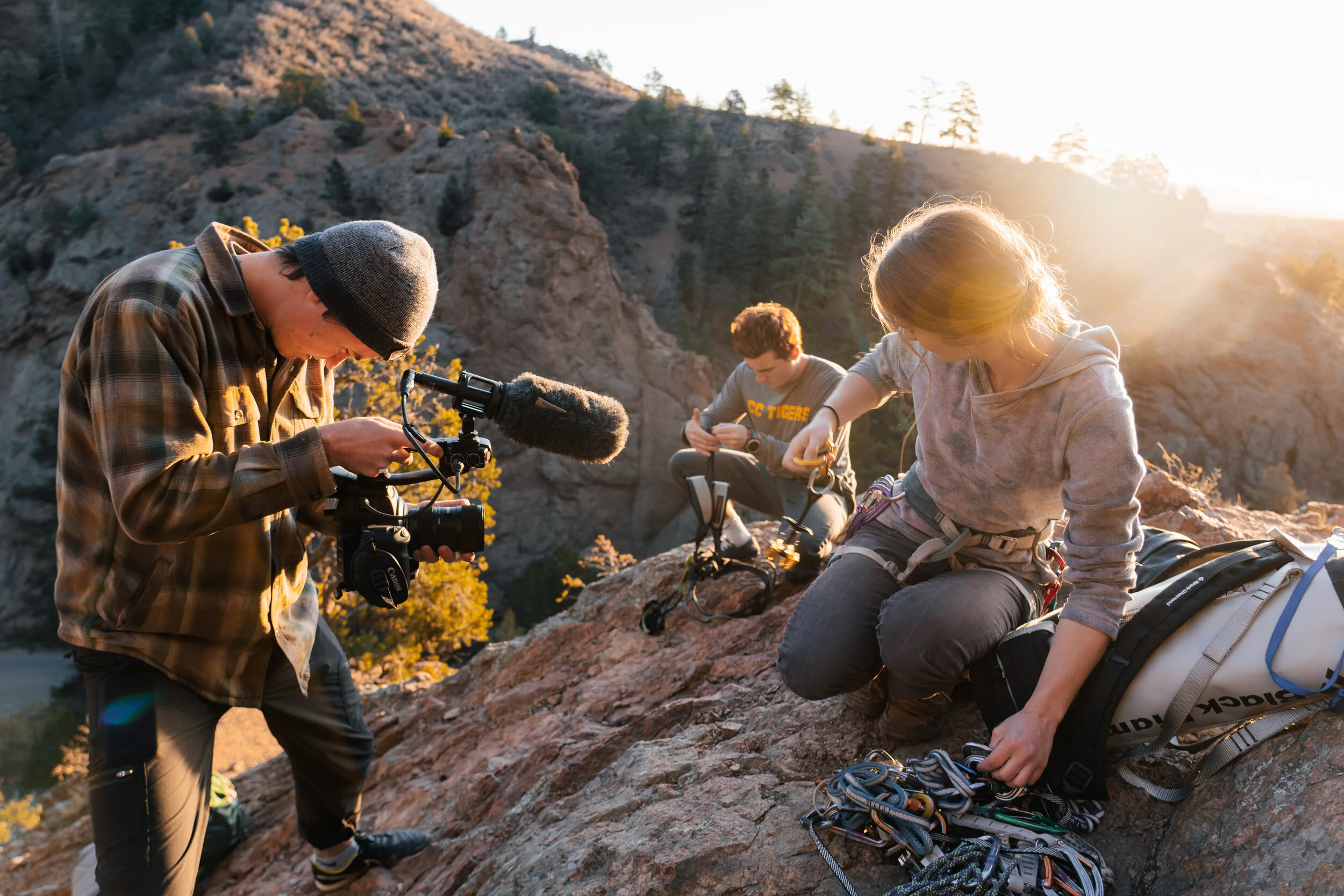 Three people preparing climbing gear on a rocky hill at sunset, with mountains and trees in the background.