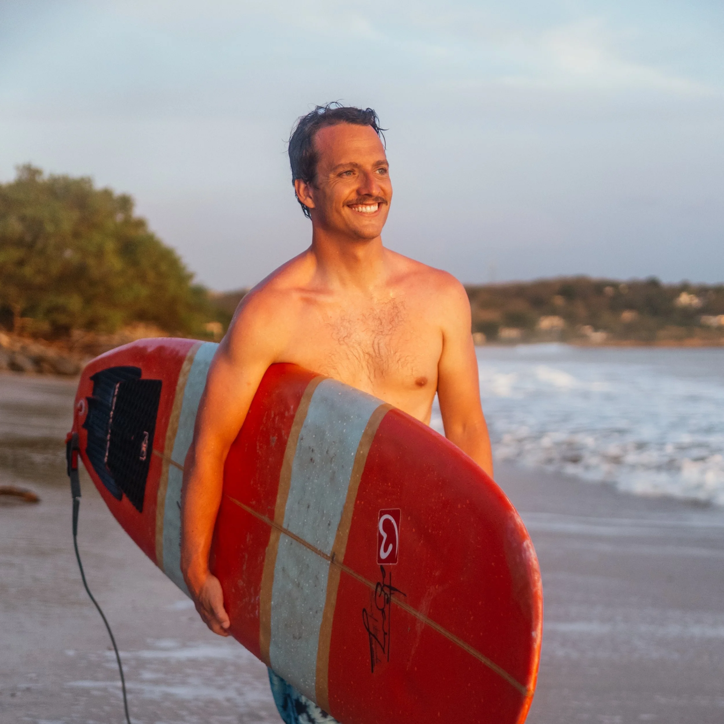 A man with a mustache, flying a red and beige surfboard, stands on the beach with the ocean and a tree-lined shoreline in the background, smiling.