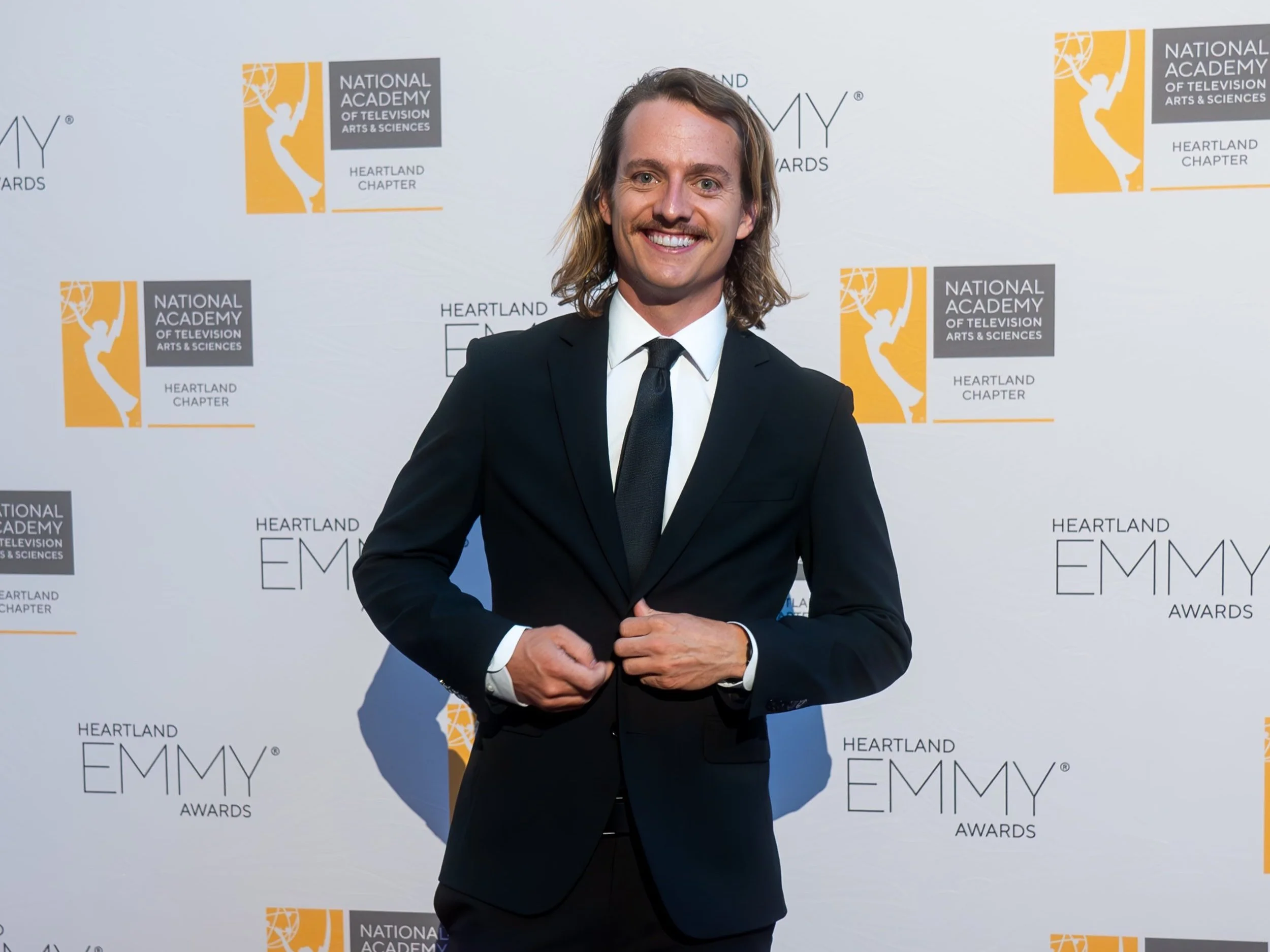 A man in a black suit, white shirt, and black tie standing on the red carpet at the Heartland Emmy Awards, smiling at the camera with a white background featuring logos of the National Academy of Television Arts & Sciences and Heartland Chapter.