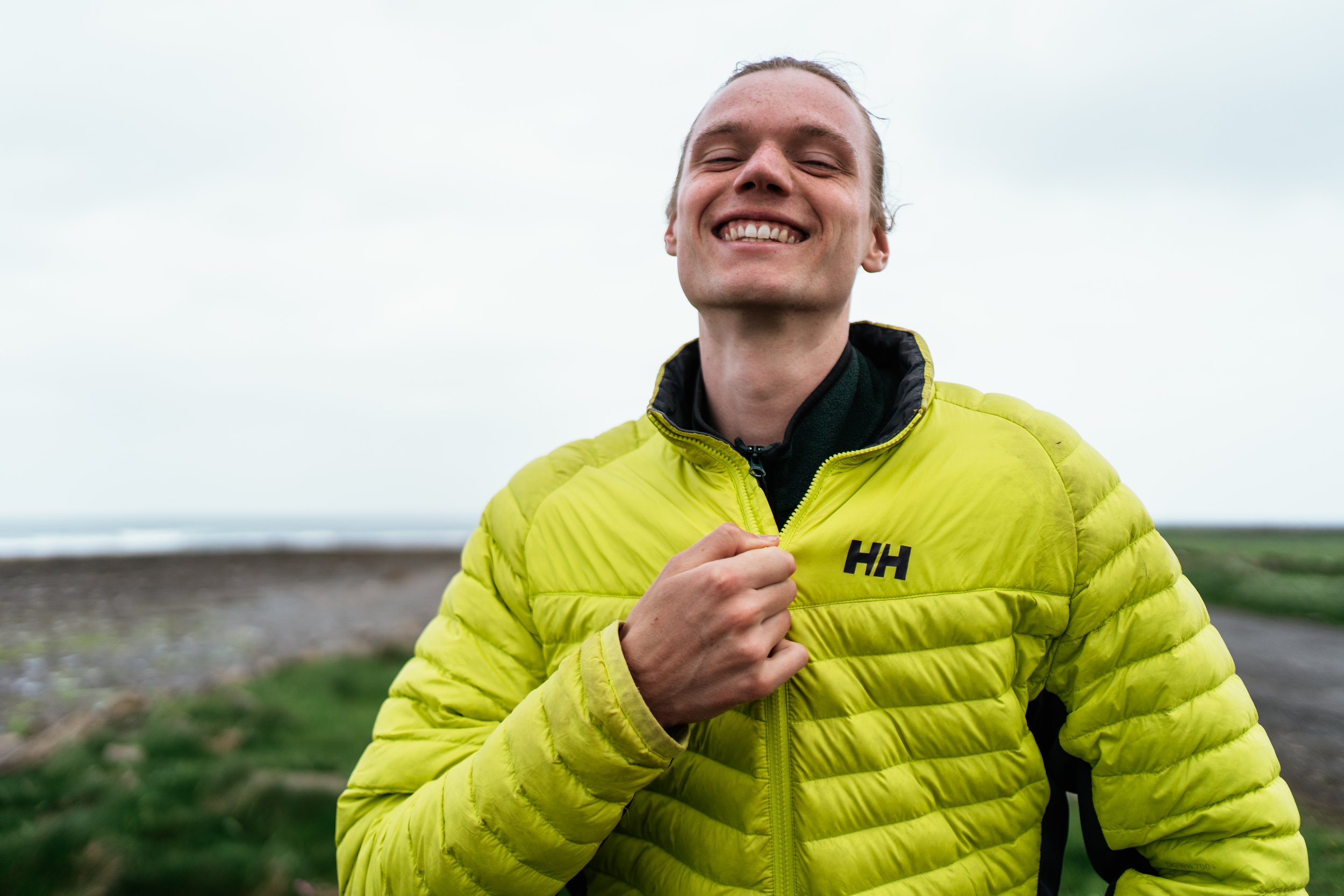 Smiling man wearing a bright yellow puffy jacket outdoors on a cloudy day.