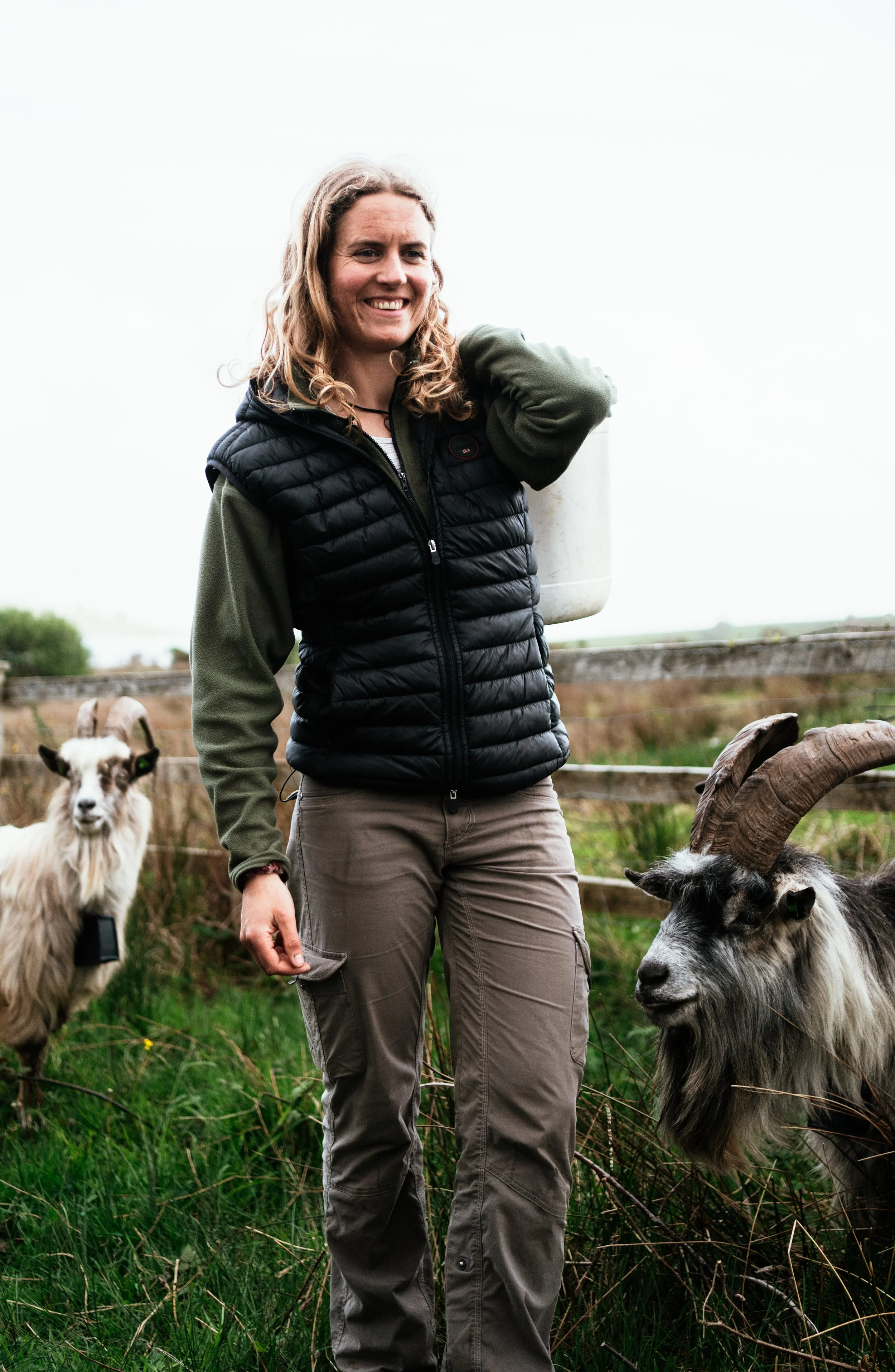 A woman smiling outdoors around goats in a grassy area, with wooden fencing in the background.