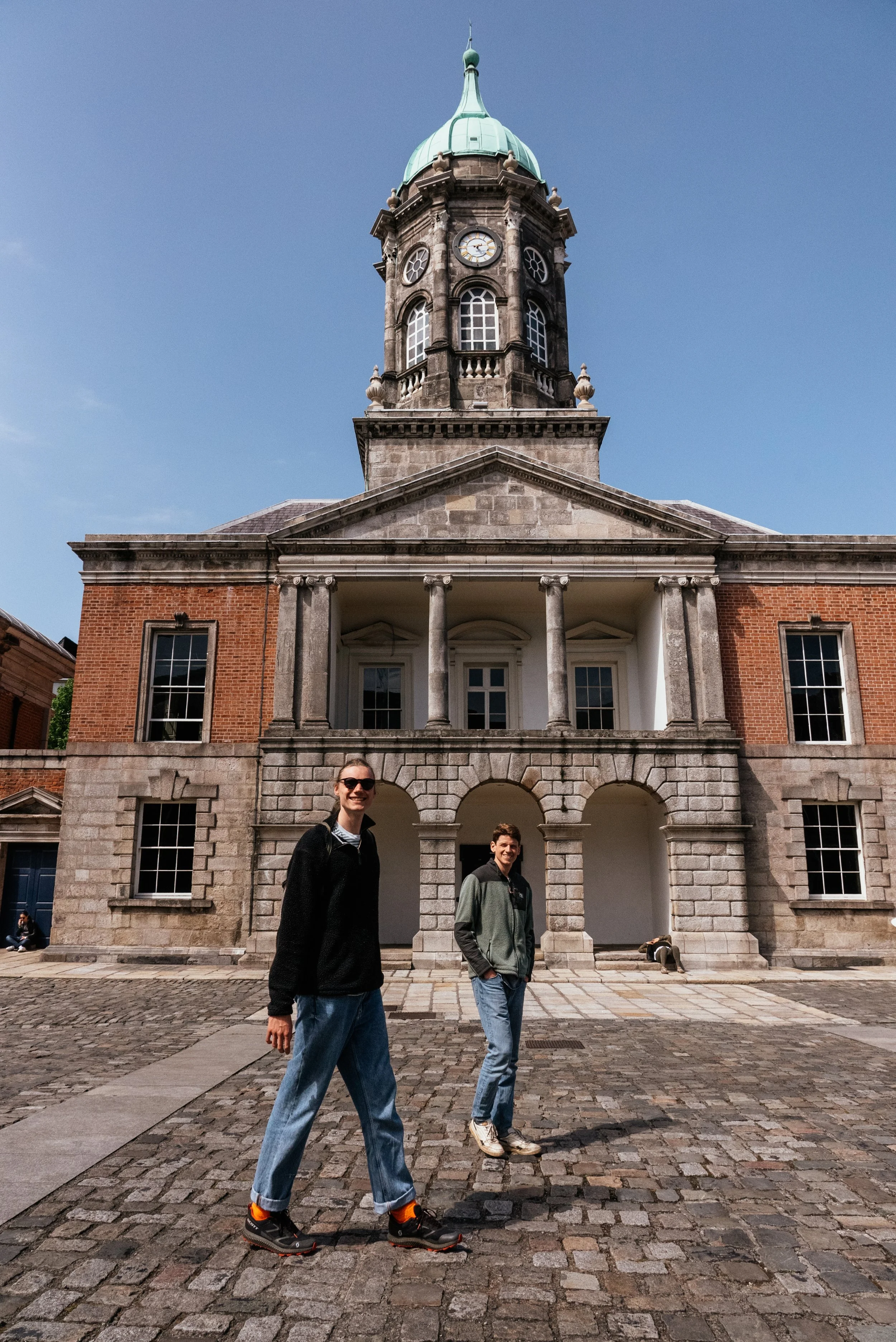 Two young people standing on a cobblestone street in front of a historic building with a clock tower, brick and stone facade, and tall windows, under a blue sky.