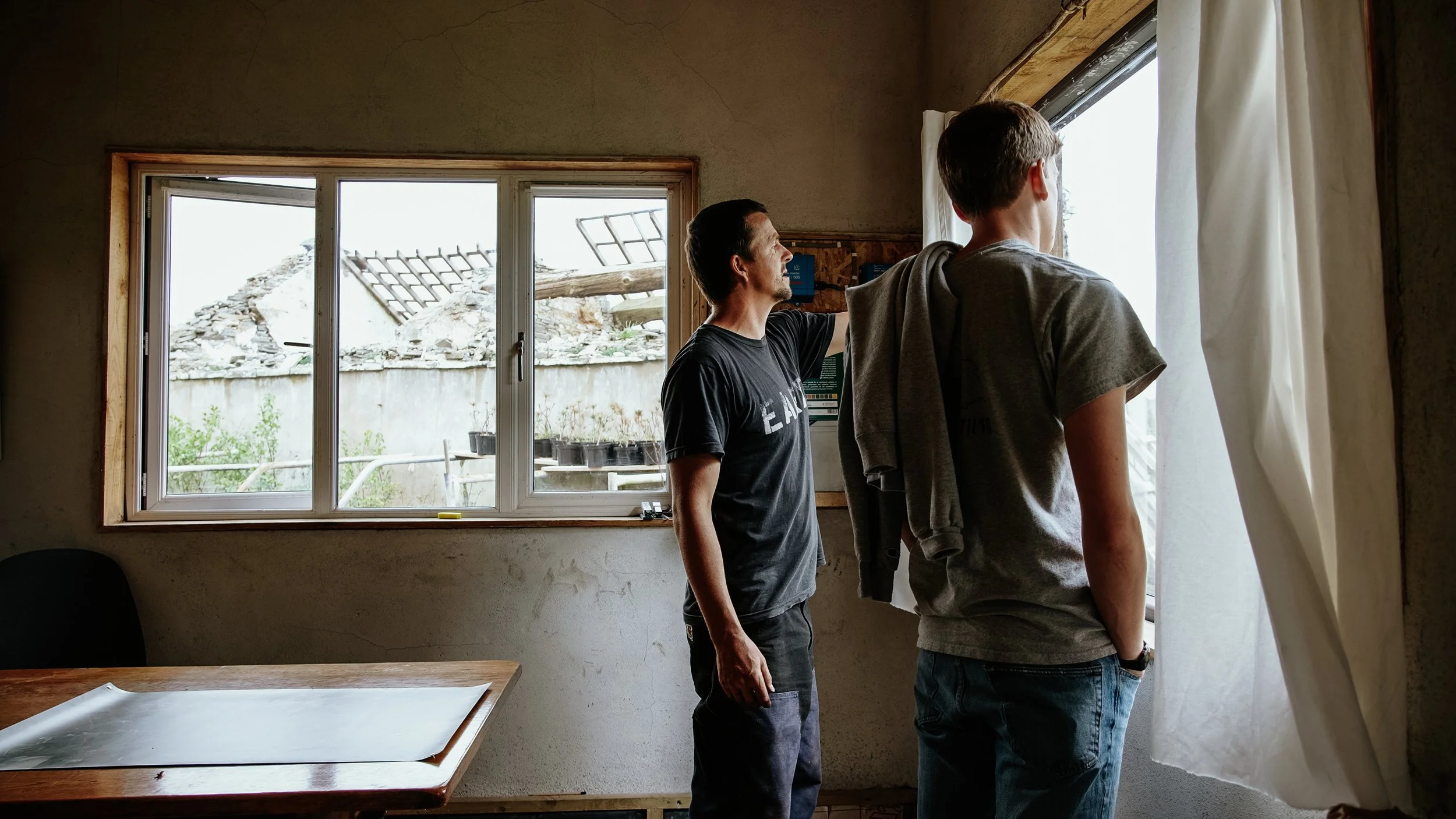 Two men standing inside a room looking out of a large window with curtains. One man is touching the window frame, and the other man is looking outside. The room has a wooden table and a chair. Outside the window, there is damage to the roof and some 