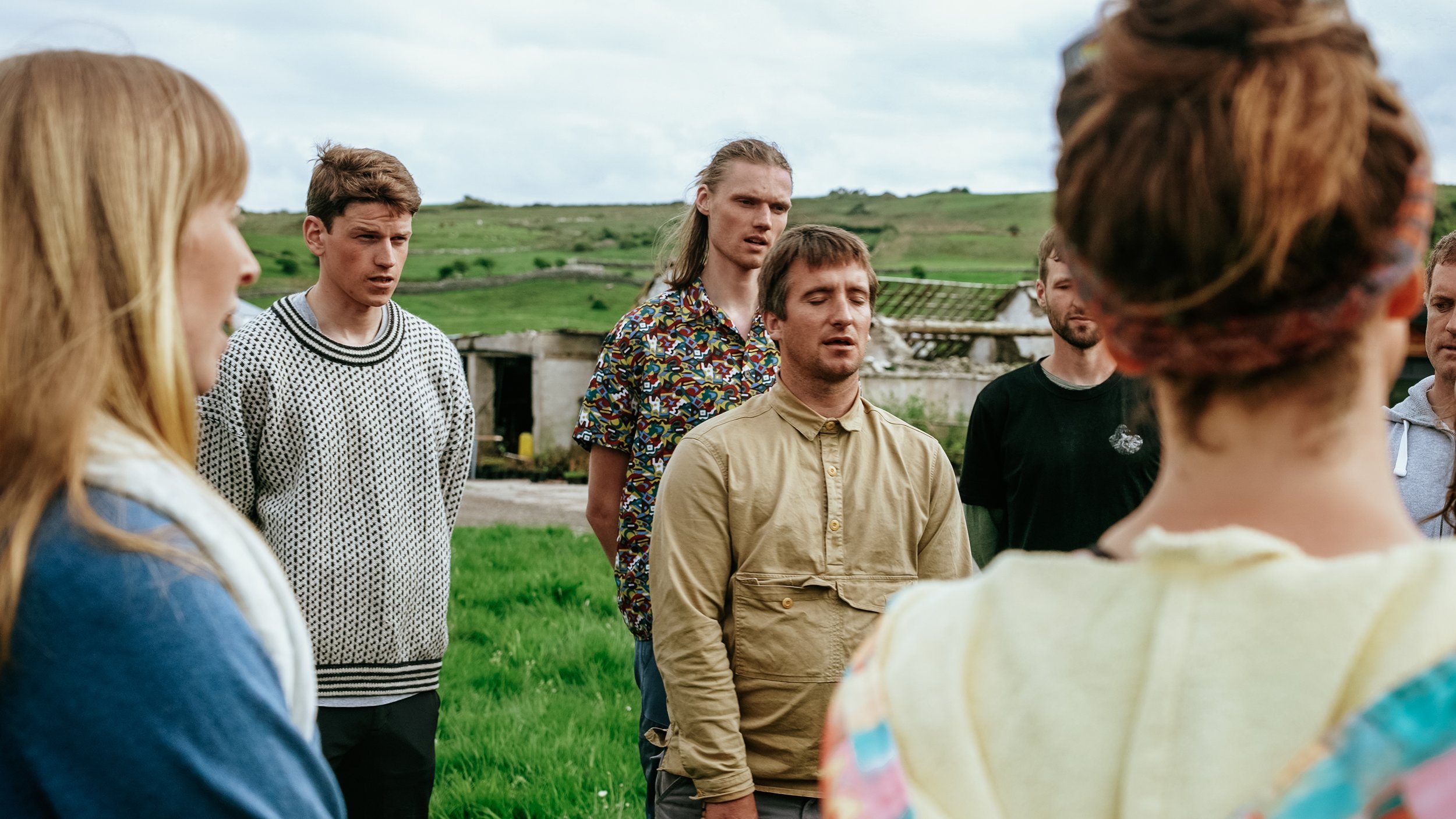 Group of young adults standing outdoors on a grassy field, appearing to be in a serious discussion or moment of reflection.