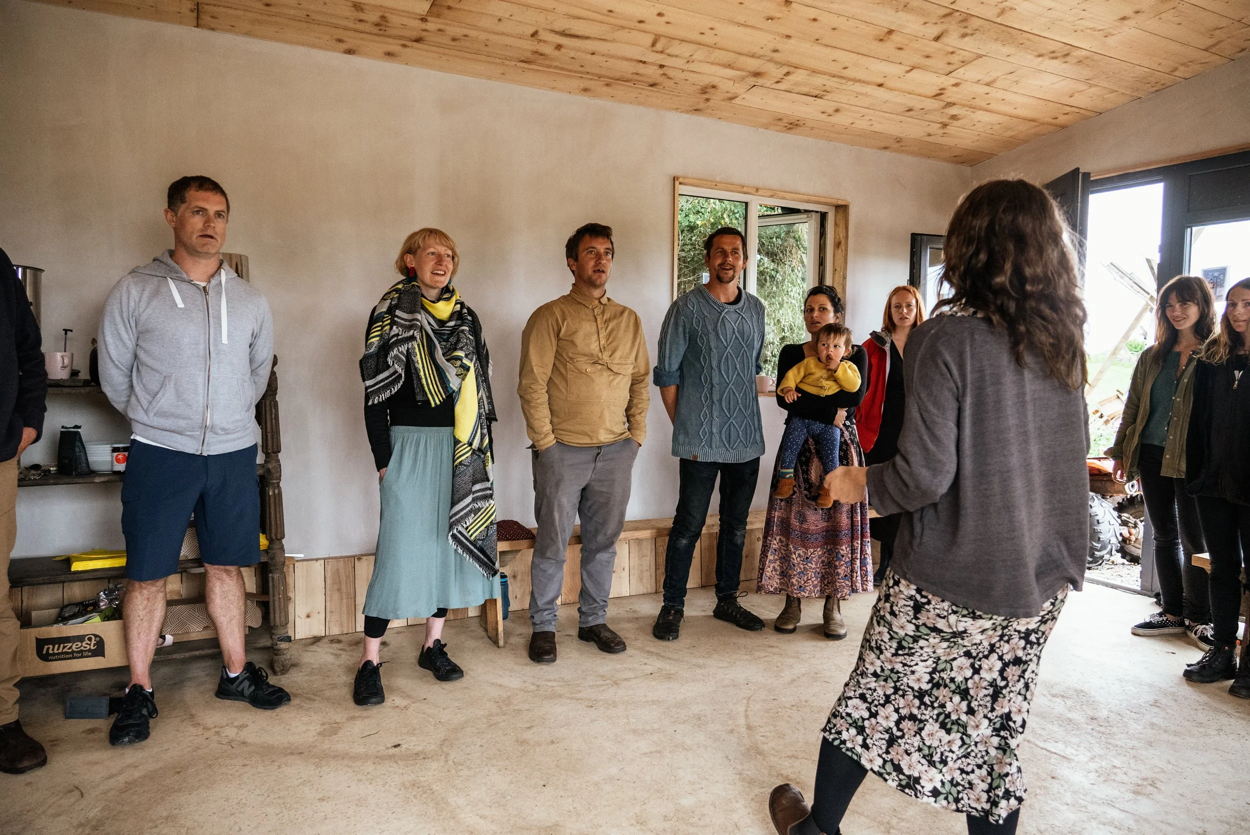 A woman with curly hair, dressed in a gray blazer and floral skirt, appears to be leading a group of people in a room with natural light, wooden ceiling, and beige walls. The group is listening attentively, with some smiling and others looking focuse