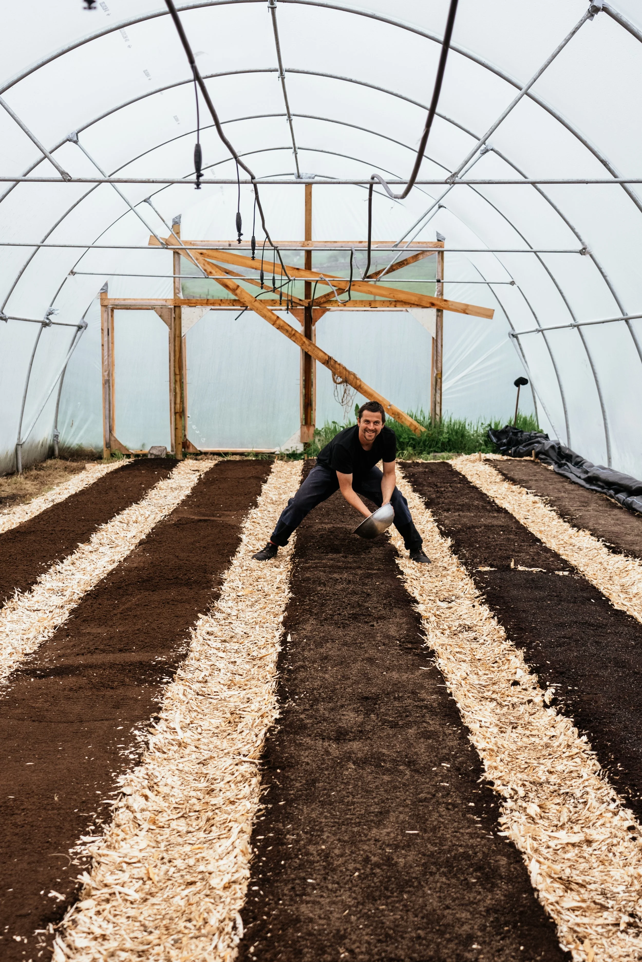 A man planting seeds inside a greenhouse with rows of soil and mulch, smiling at the camera.