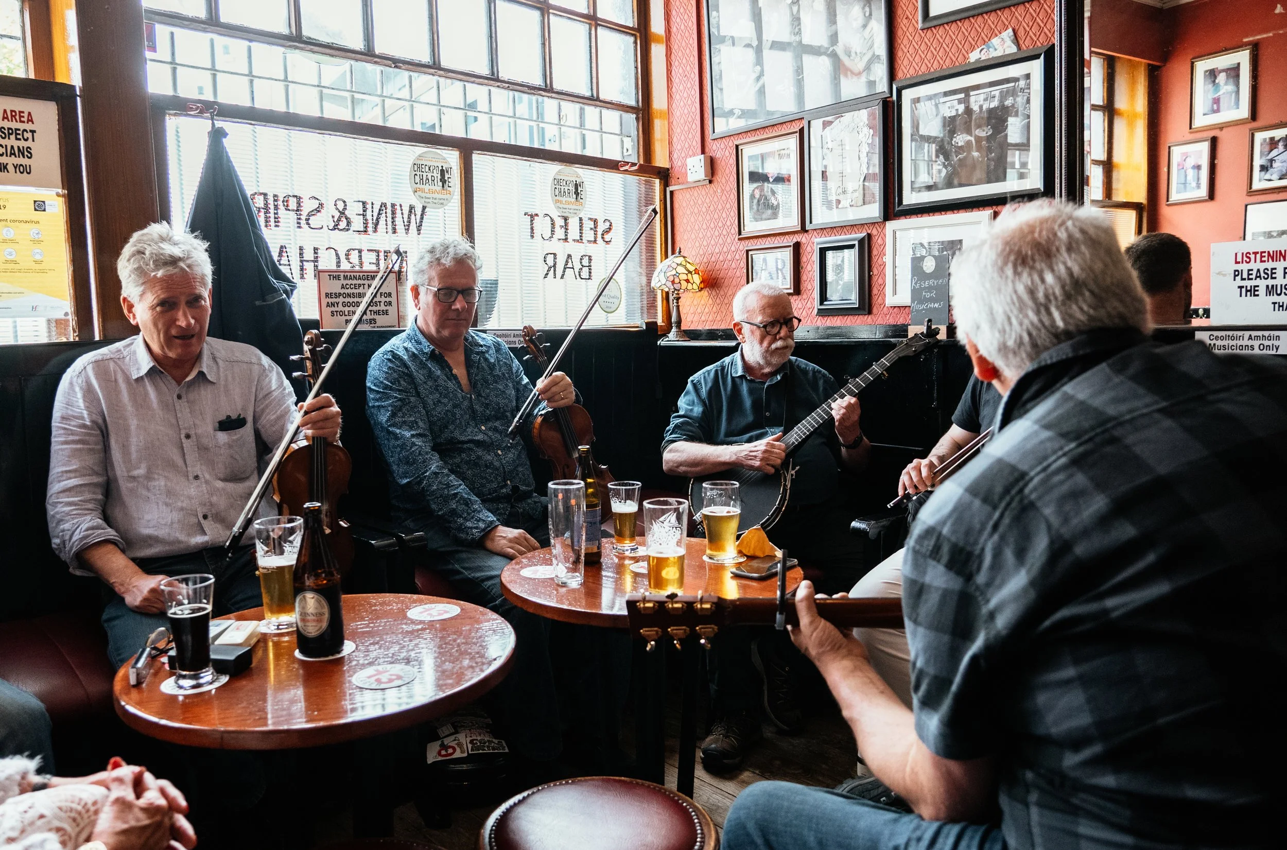 A group of musicians playing instruments in a small pub or cafe, including violins, guitar, and a ukulele, with several glasses of beer on the table