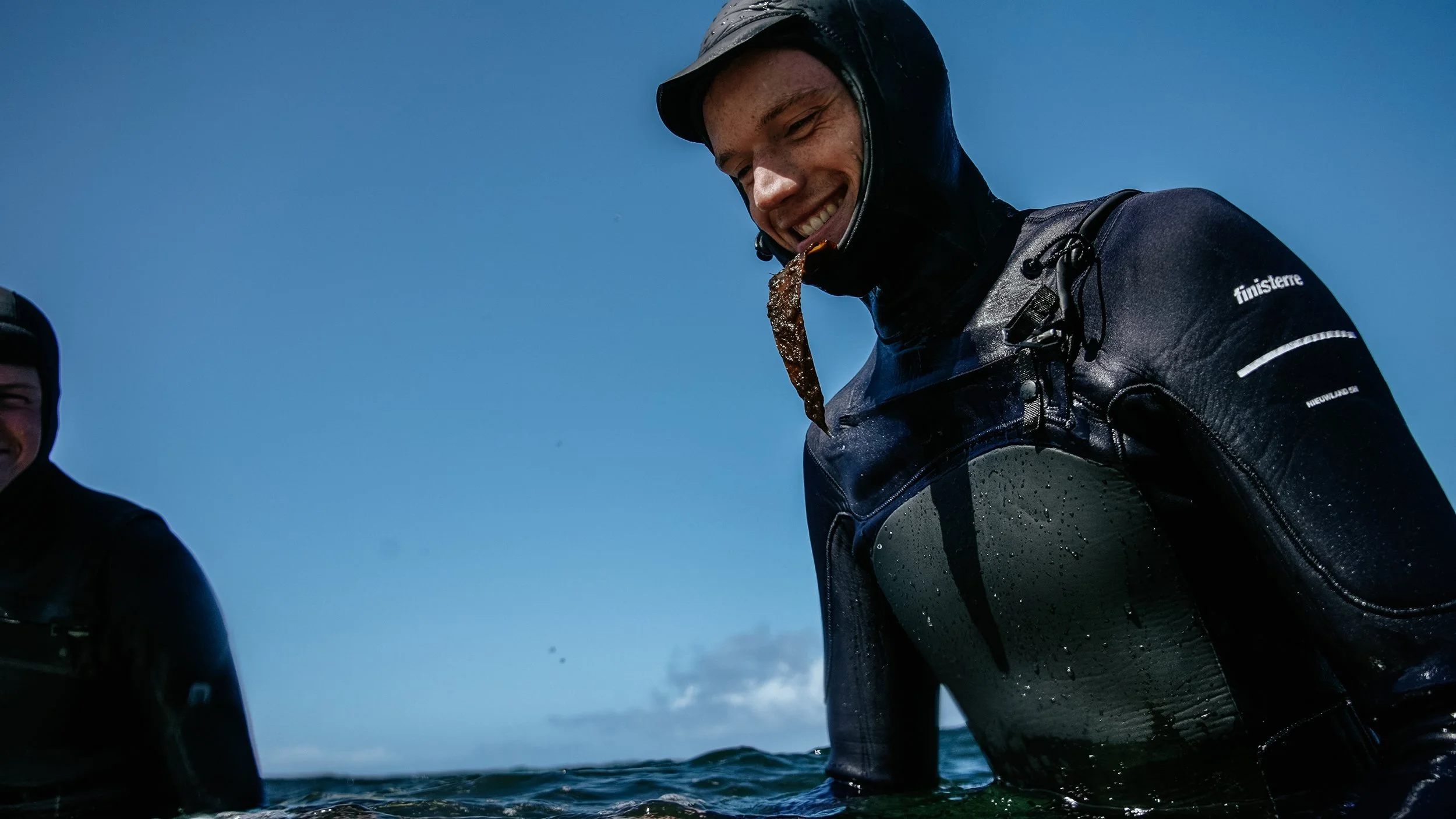 Man in a wetsuit smiling, holding seaweed in his mouth, standing in the ocean against a cloudy sky.