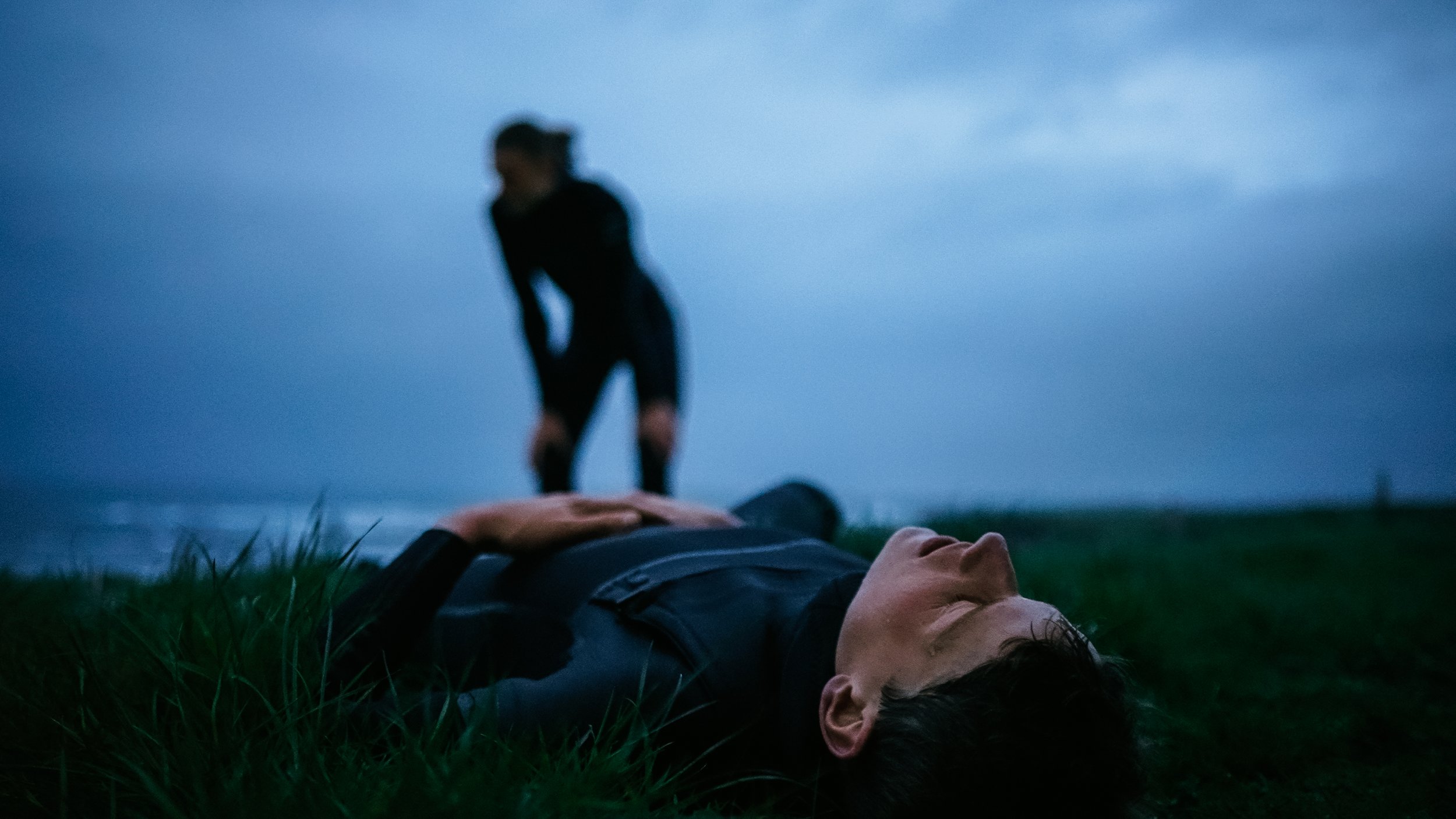A person lying on the ground with closed eyes in grass, another person blurred in the background near water during dusk or dawn.