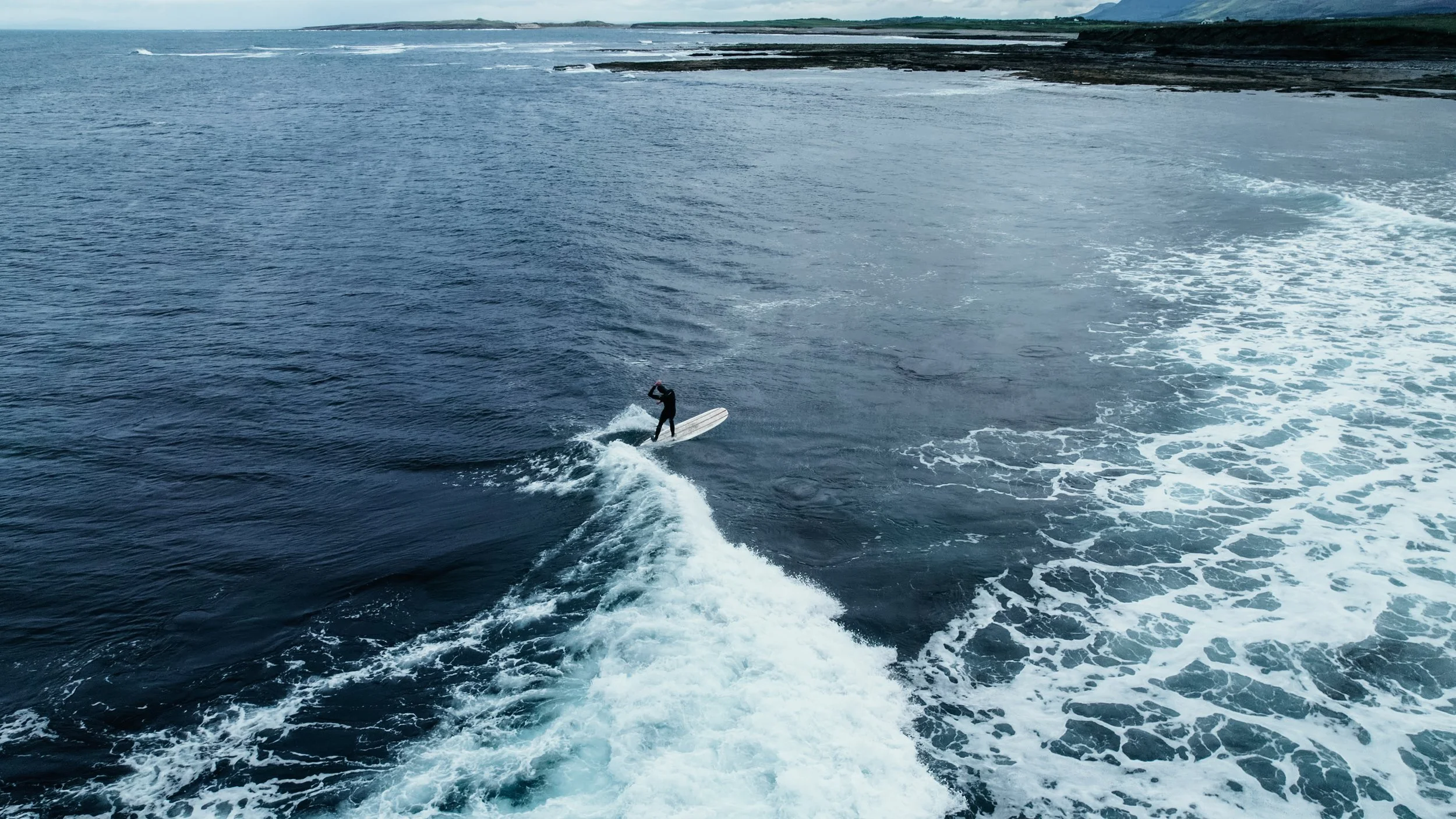 A person wearing a wetsuit surfing on a white surfboard on the ocean, with the waves and shoreline in the background.