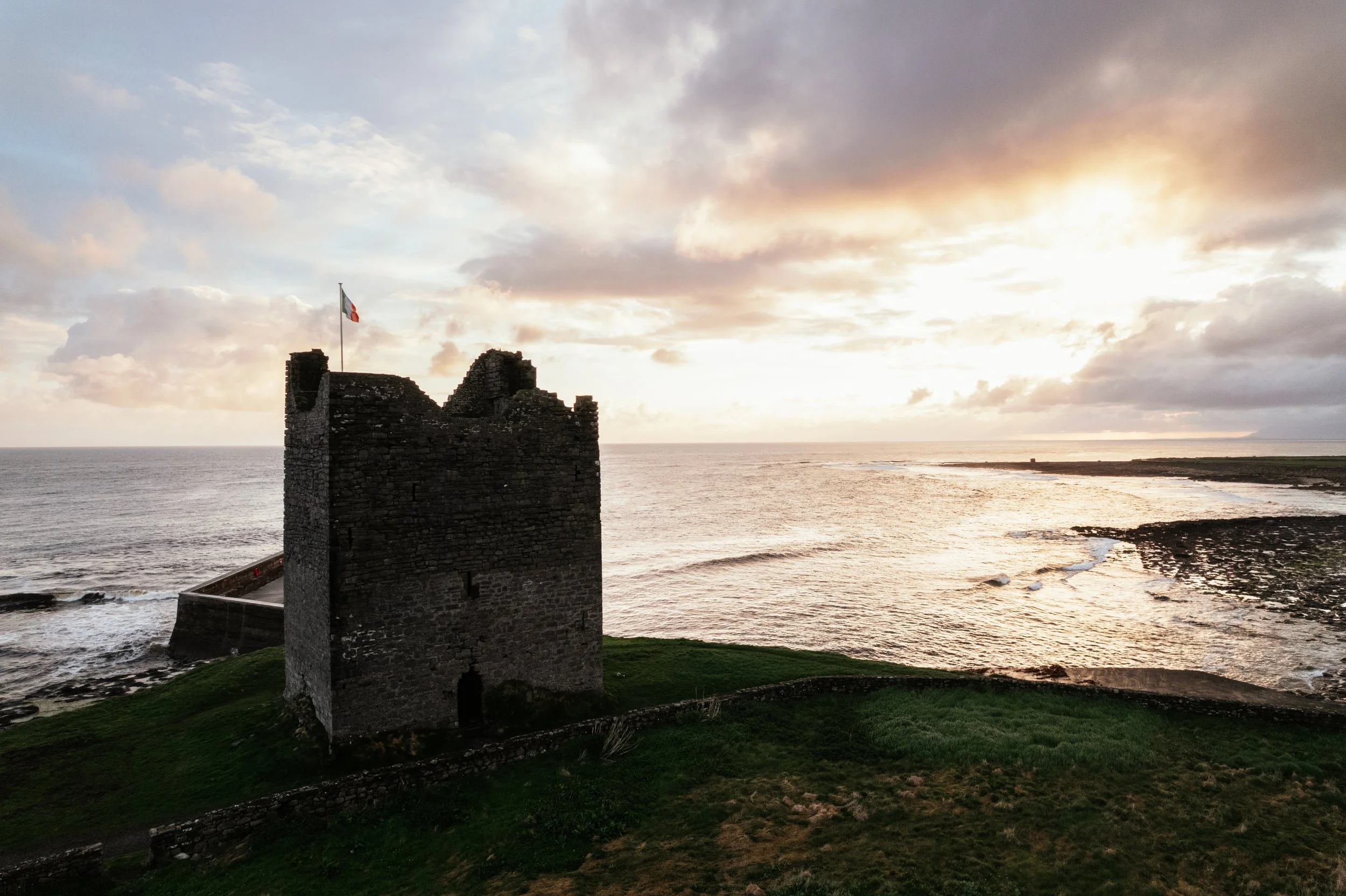 An old stone tower with a flag on top, overlooking the ocean at sunset, with a cloudy sky and grassy area in the foreground.