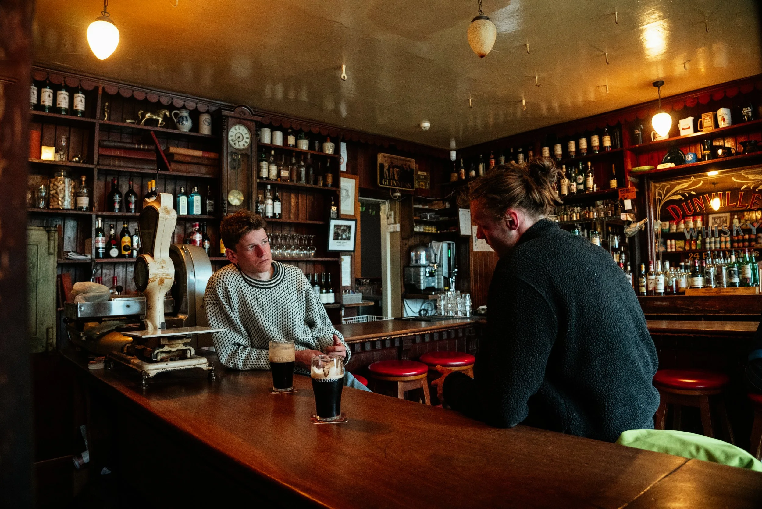 Two young men sitting at a bar, engaged in conversation, with drinks in front of them, inside a dimly lit pub with shelves of liquor bottles behind the bar.