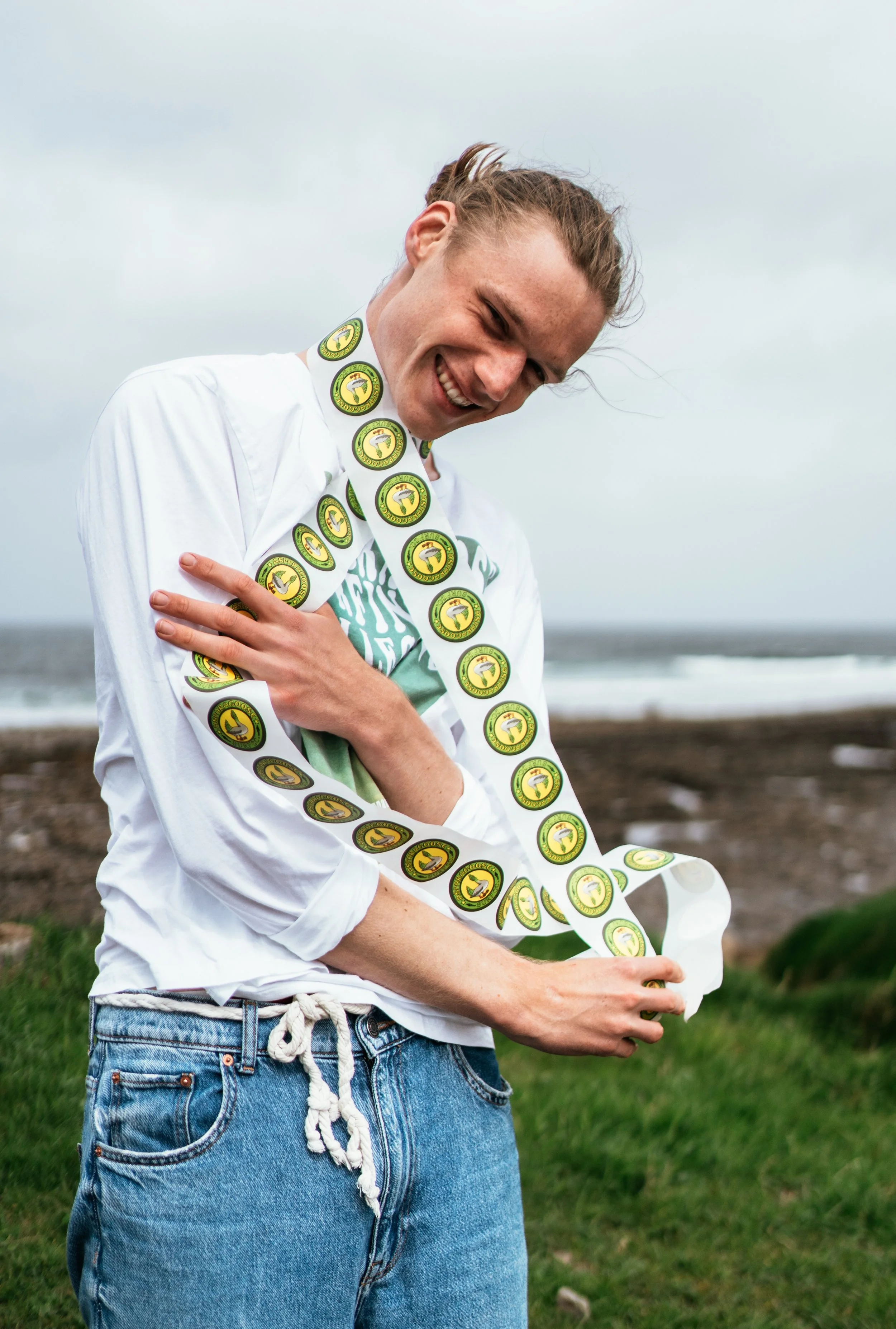 A smiling young woman with a long striped necktie made of circular yellow and green patches, standing outdoors near the beach with grass and sand in the background.