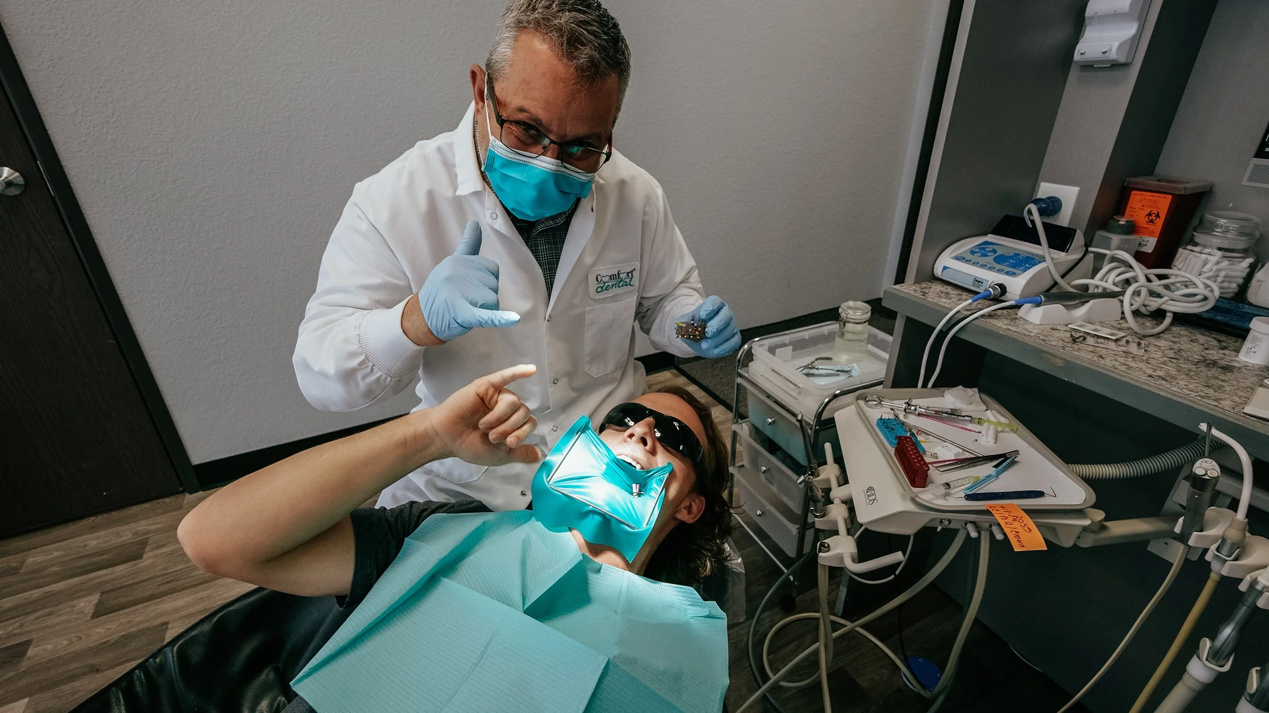 A dentist in a white coat and mask with protective glasses works on a patient in a dental chair, who is smiling and wearing protective eyewear, in a dental office with dental tools and equipment around.