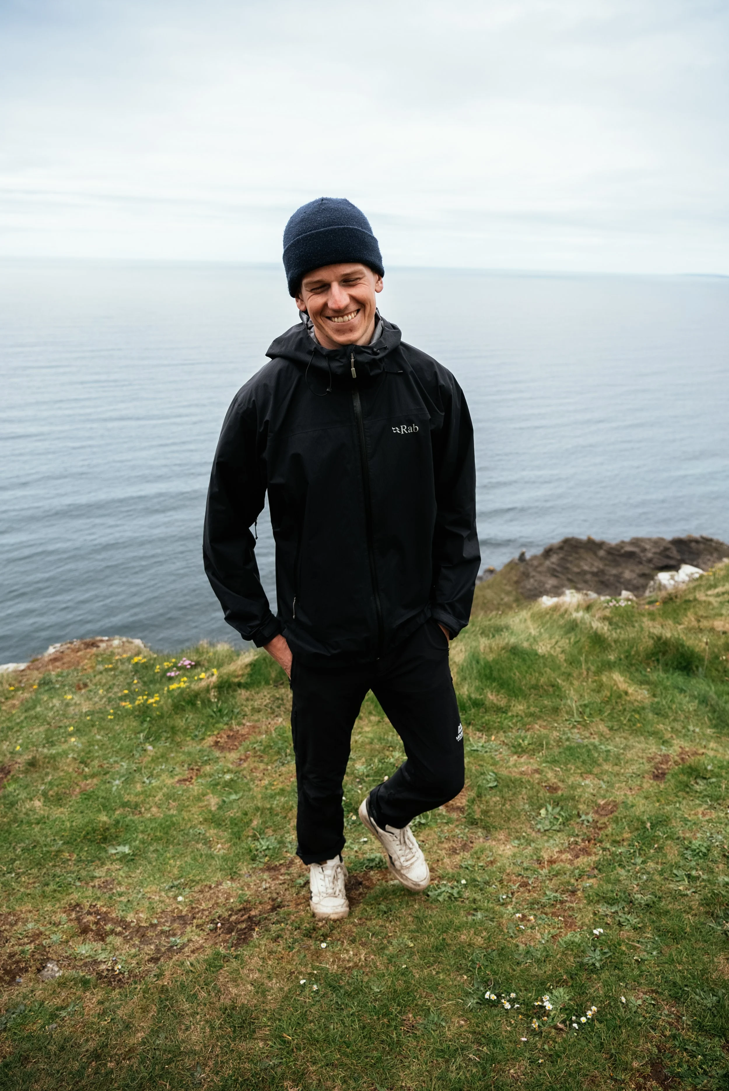 A young man smiling, dressed in black raincoat and pants, wearing a navy beanie, standing on grassy cliff with rocky coastline and ocean in the background under cloudy sky.
