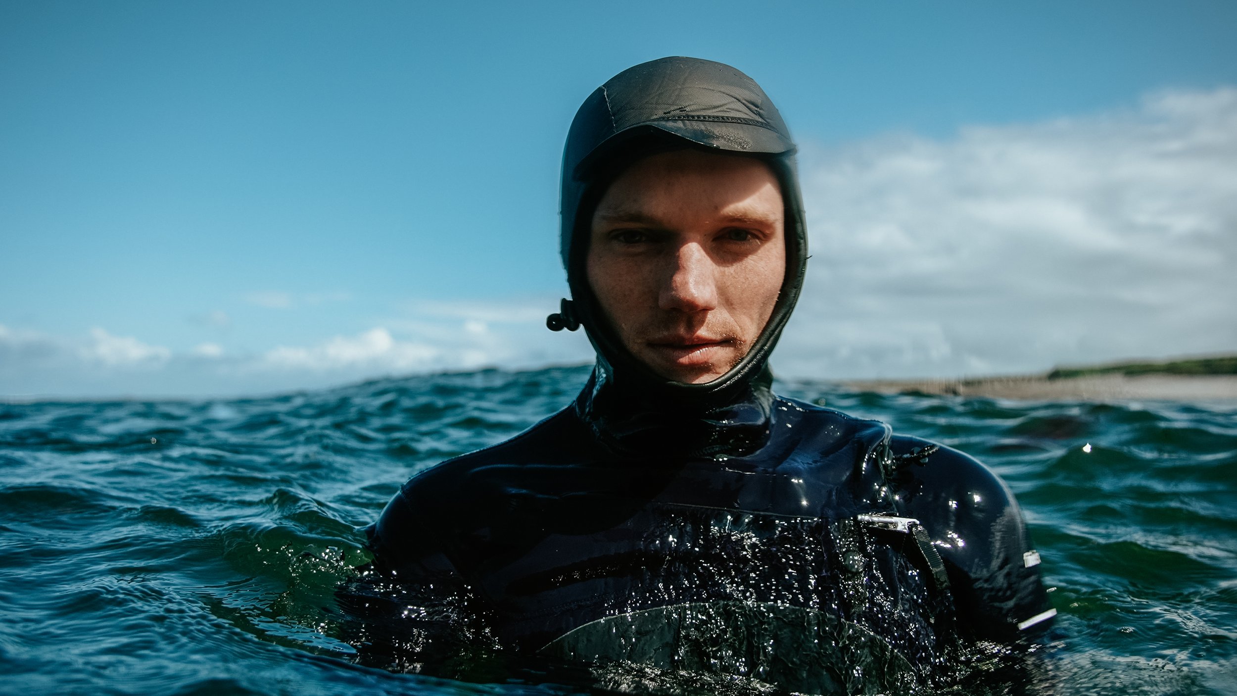 A young man in a black wetsuit and helmet stands in the ocean with a cloudy sky behind him.