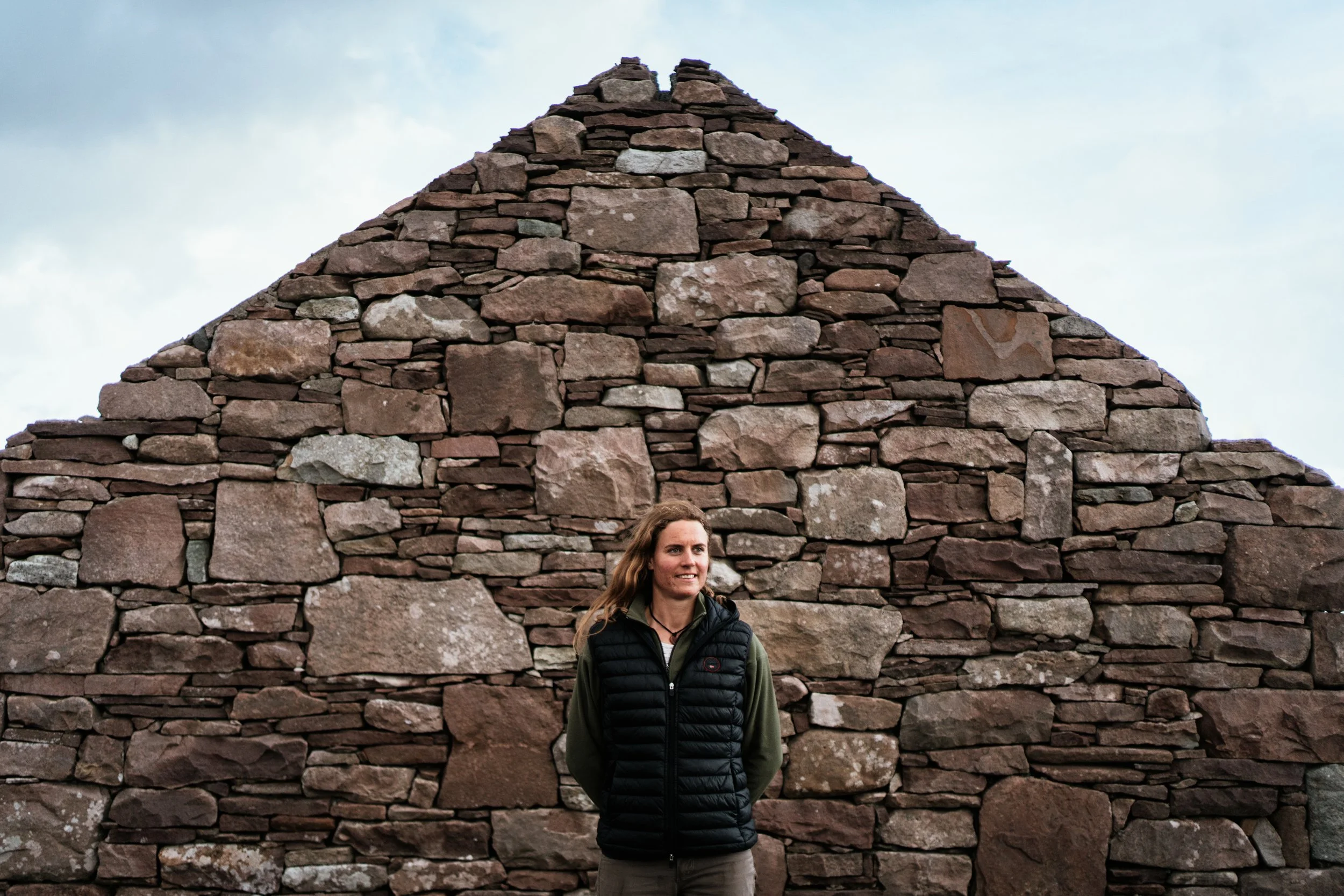 A woman standing in front of a large ancient stone wall, smiling, wearing a black puffer vest and green long-sleeve shirt.