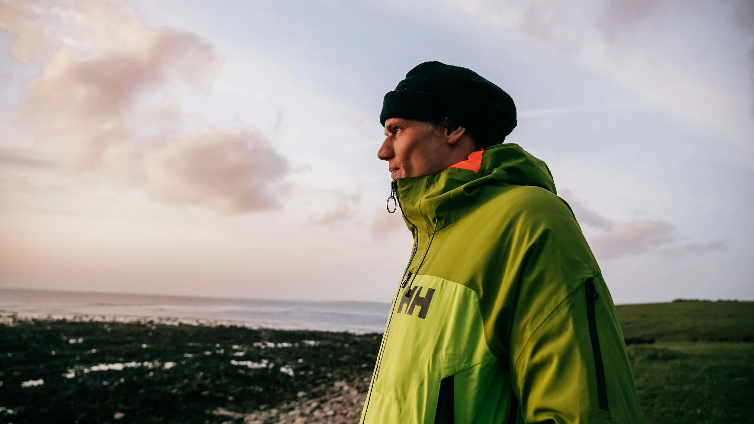 A man wearing a black beanie and a green jacket standing outdoors near a coastline with clouds in the sky.