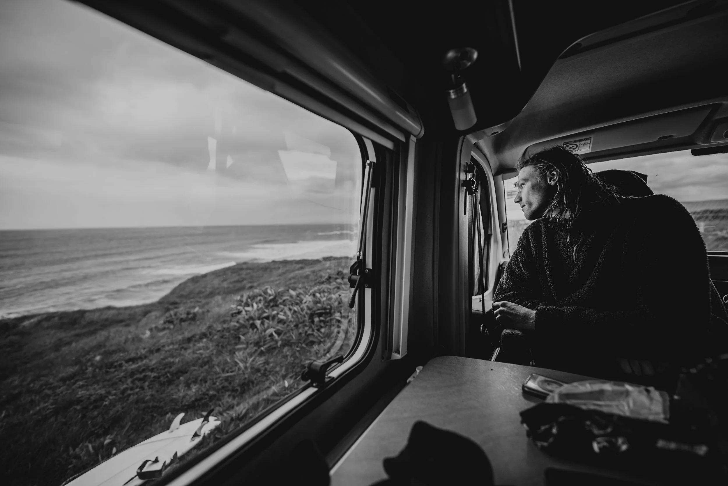 A person with long hair looking out the window of a vehicle at the ocean, in black and white.