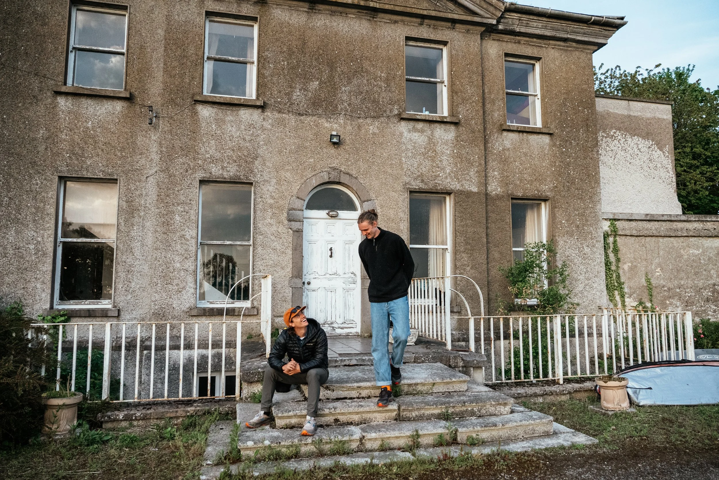 Two men conversing on the front steps of an old, weathered house with several windows, some with reflections of the sky, during daytime. The house has a white front door and a small garden area with potted plants and greenery.