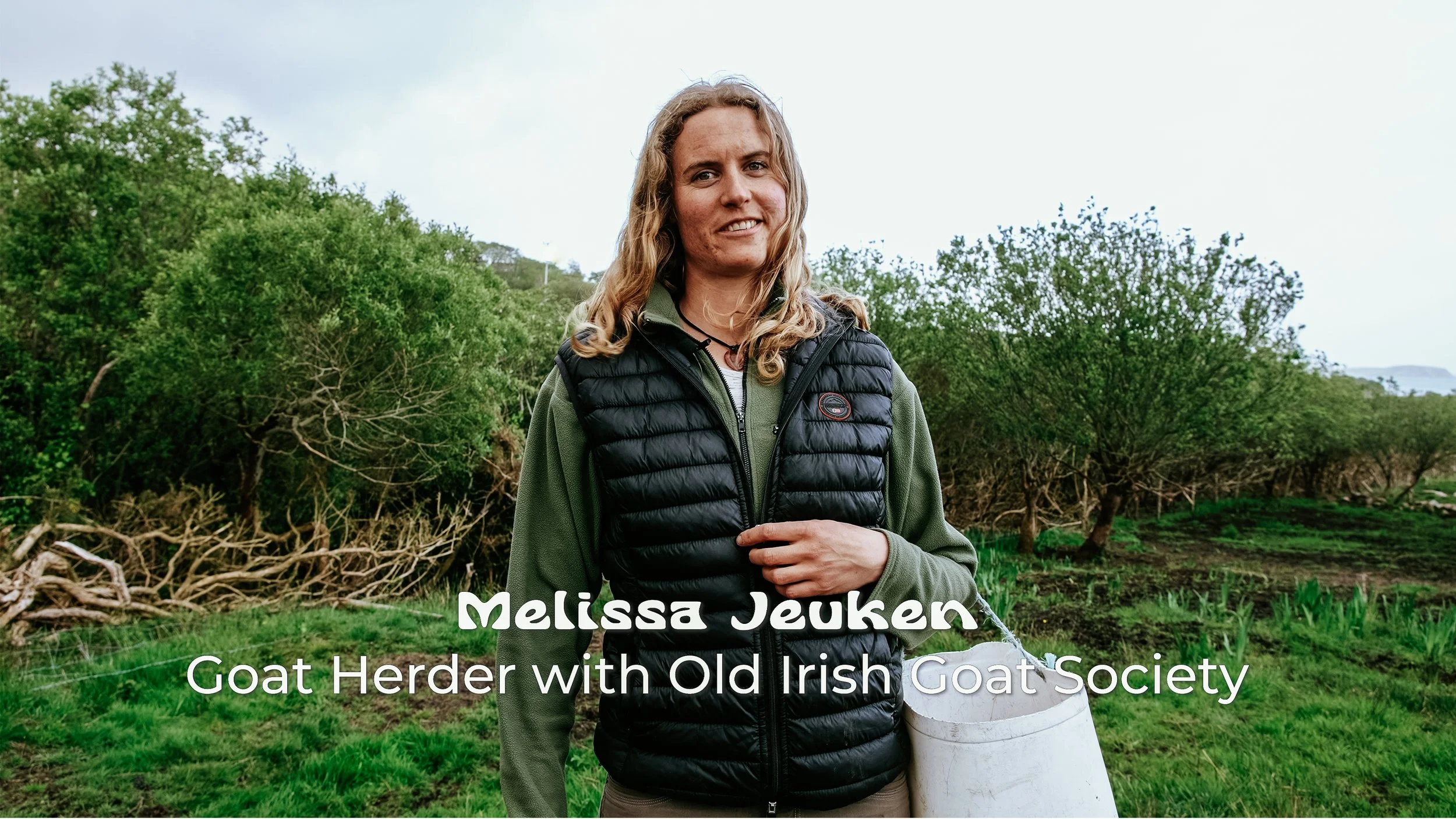 Melissa Jeuken standing outdoors in a green field with trees in the background, holding a white bucket, wearing a green jacket and black vest, smiling at the camera.