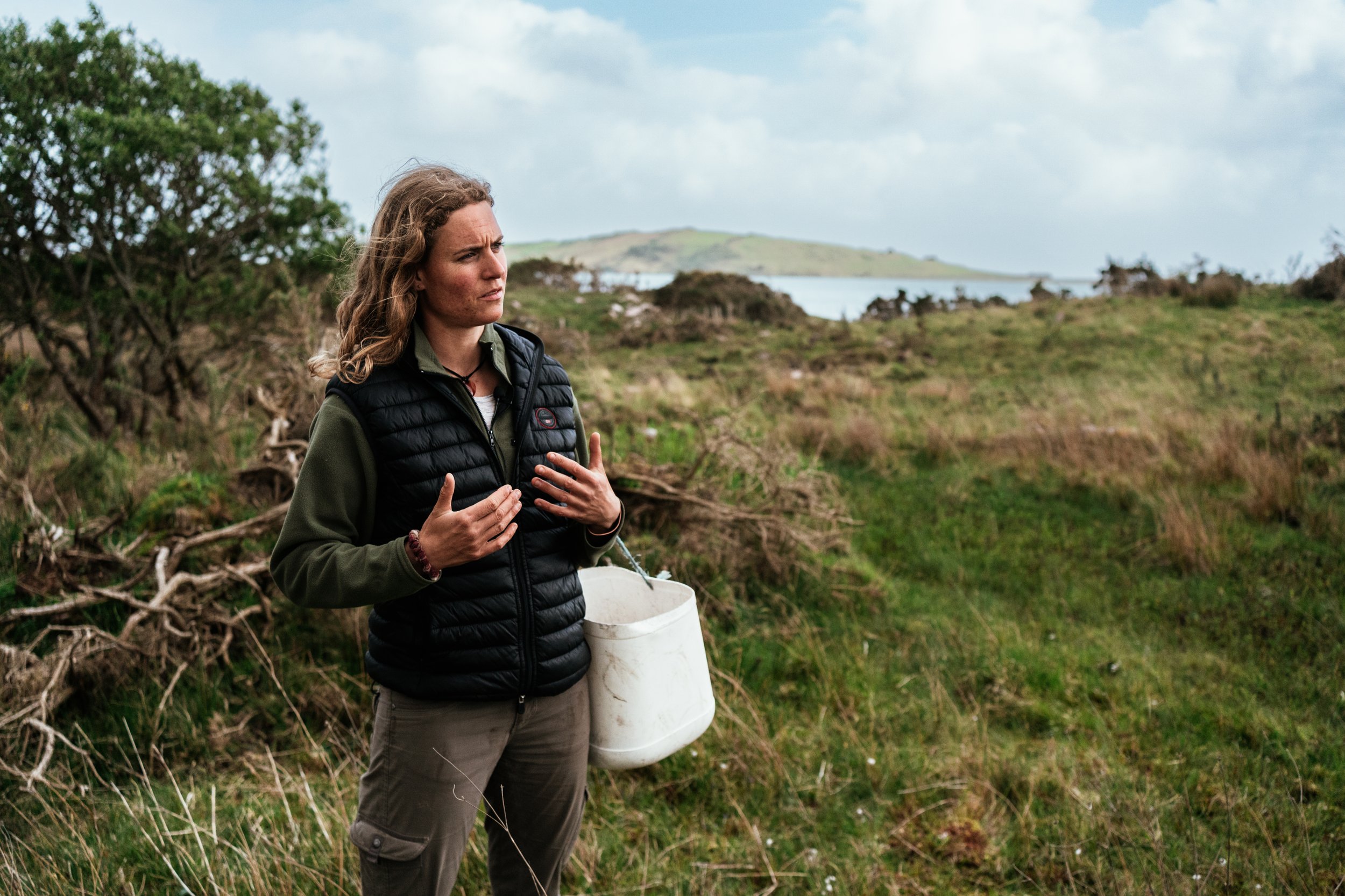 A woman standing on a grassy field near shrubs with a body of water and hills in the background, holding her hands to her chest and wearing outdoor clothing and a black puffy vest.