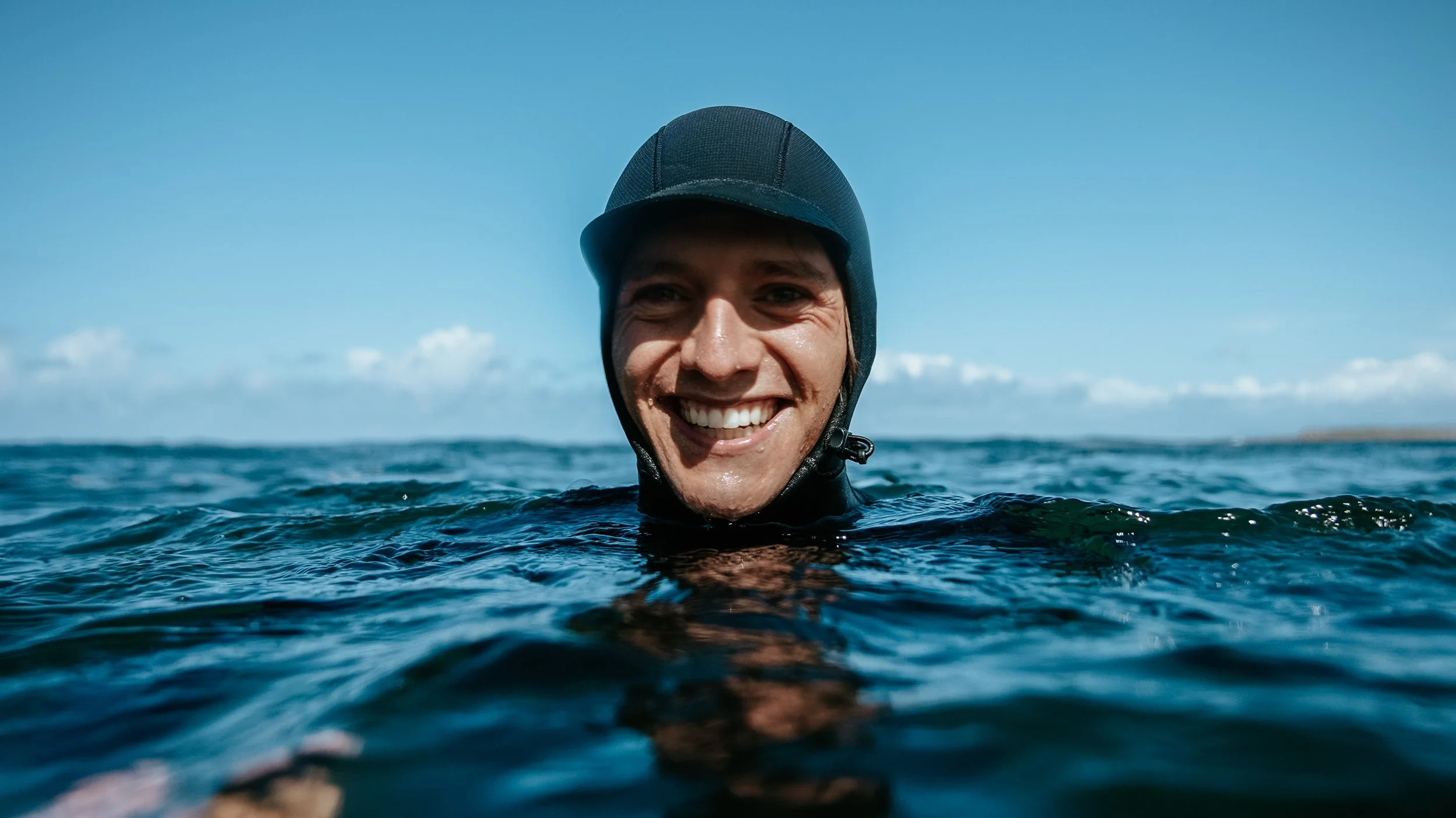 A smiling man in a wetsuit and helmet swimming in the ocean with a clear blue sky in the background.